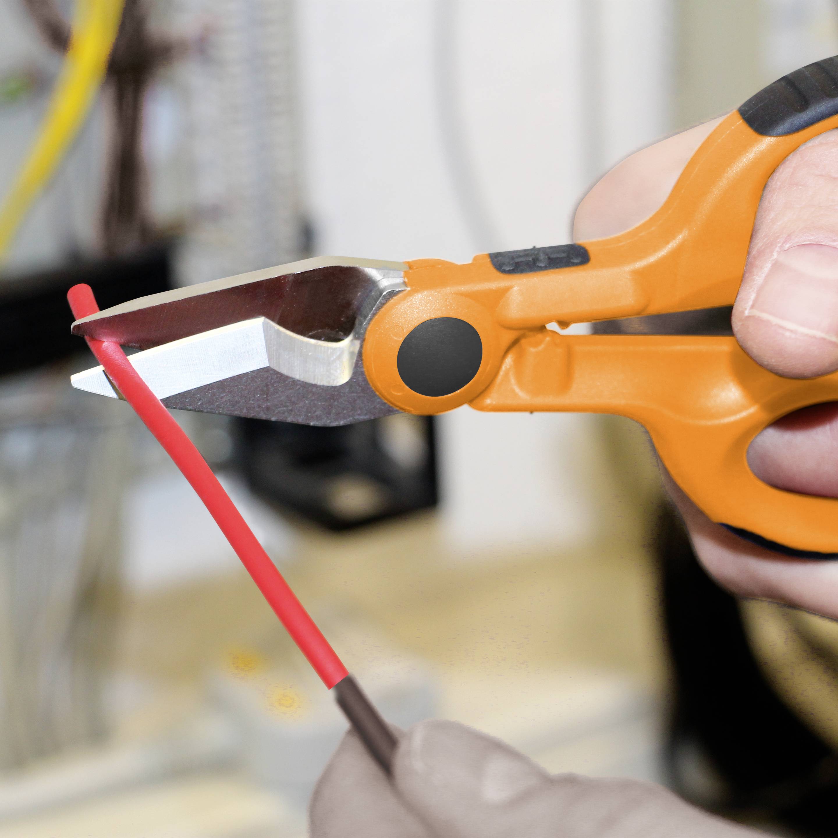 Eine Hand hält eine orangefarbene Zange und schneidet ein rotes Kabel durch. Im Hintergrund sind unscharfe technische Geräte erkennbar.