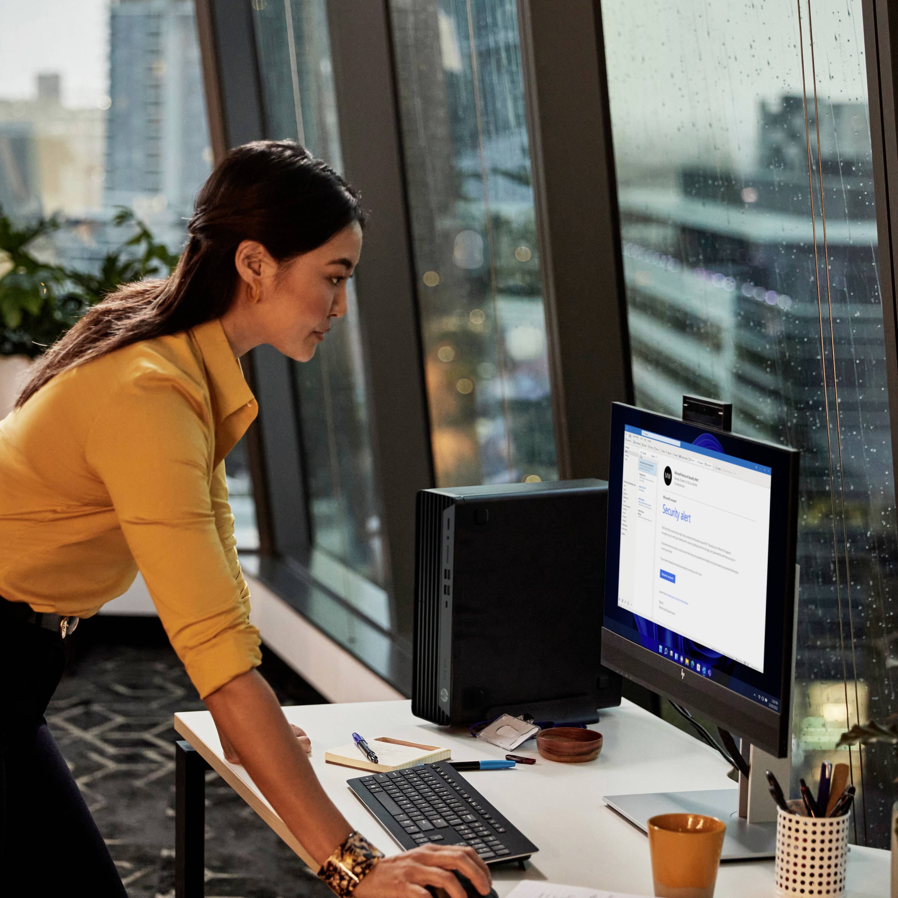 Eine Frau in einem gelben Hemd arbeitet konzentriert an einem Computer in einem modernen Büro mit großen Fenstern im Hintergrund.