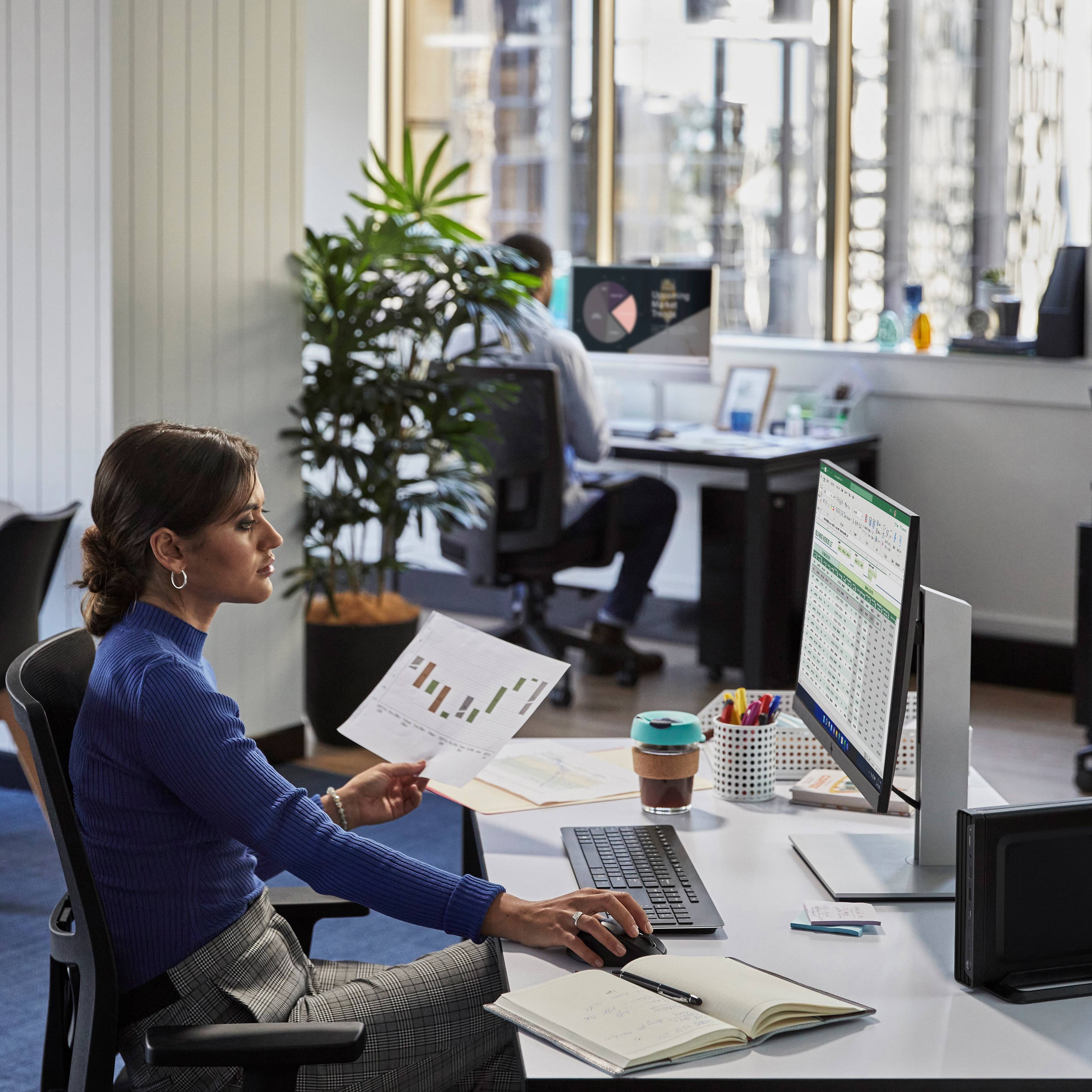 Eine Frau im Büro arbeitet am Computer mit Tabellen, hält ein Diagramm. Im Hintergrund sitzt ein Kollege. Schreibtisch mit Büroartikeln.