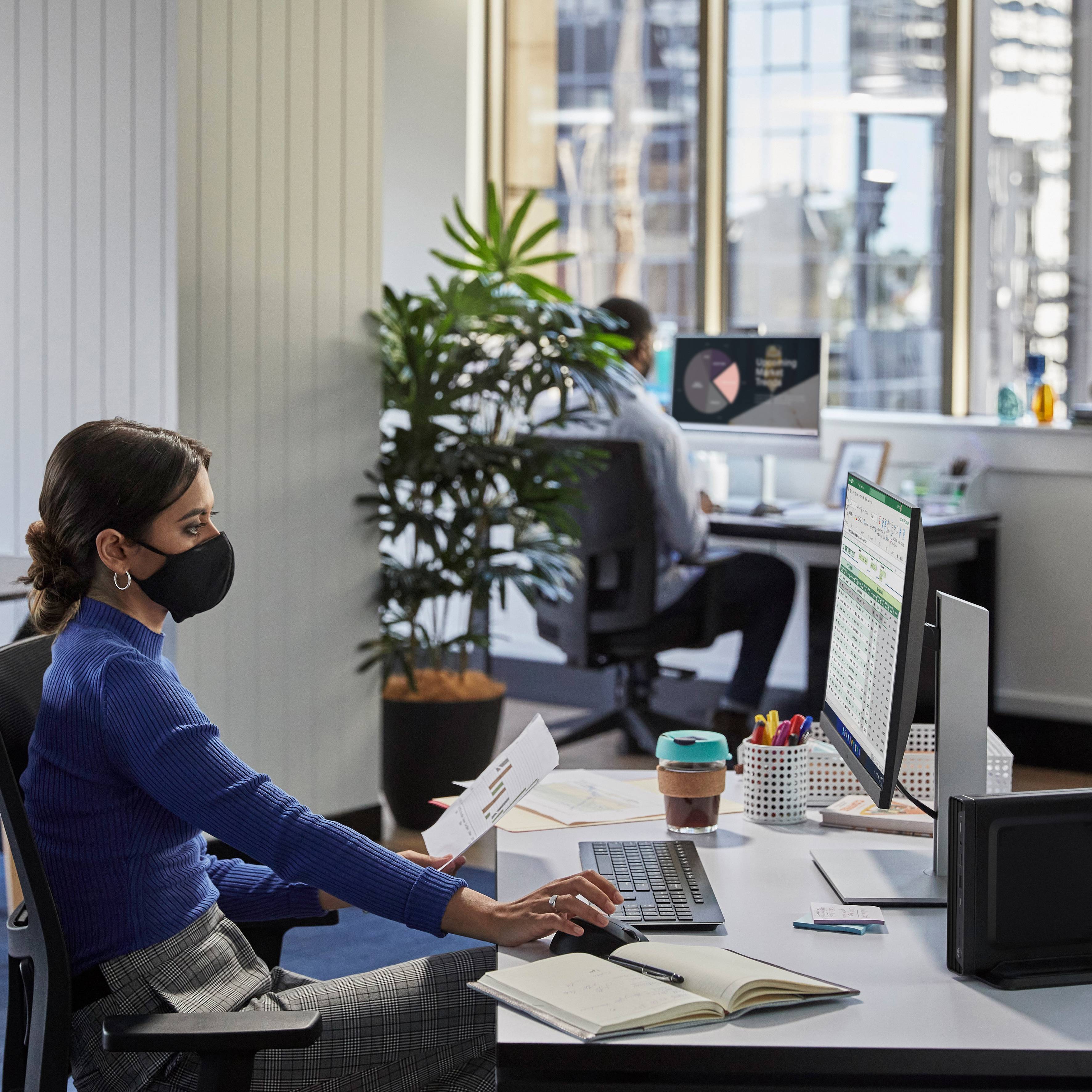 Eine Frau sitzt an einem Schreibtisch in einem Büro, prüft Dokumente. Im Hintergrund arbeitet ein Mann am Computer. Pflanzen und Fenster im Raum.