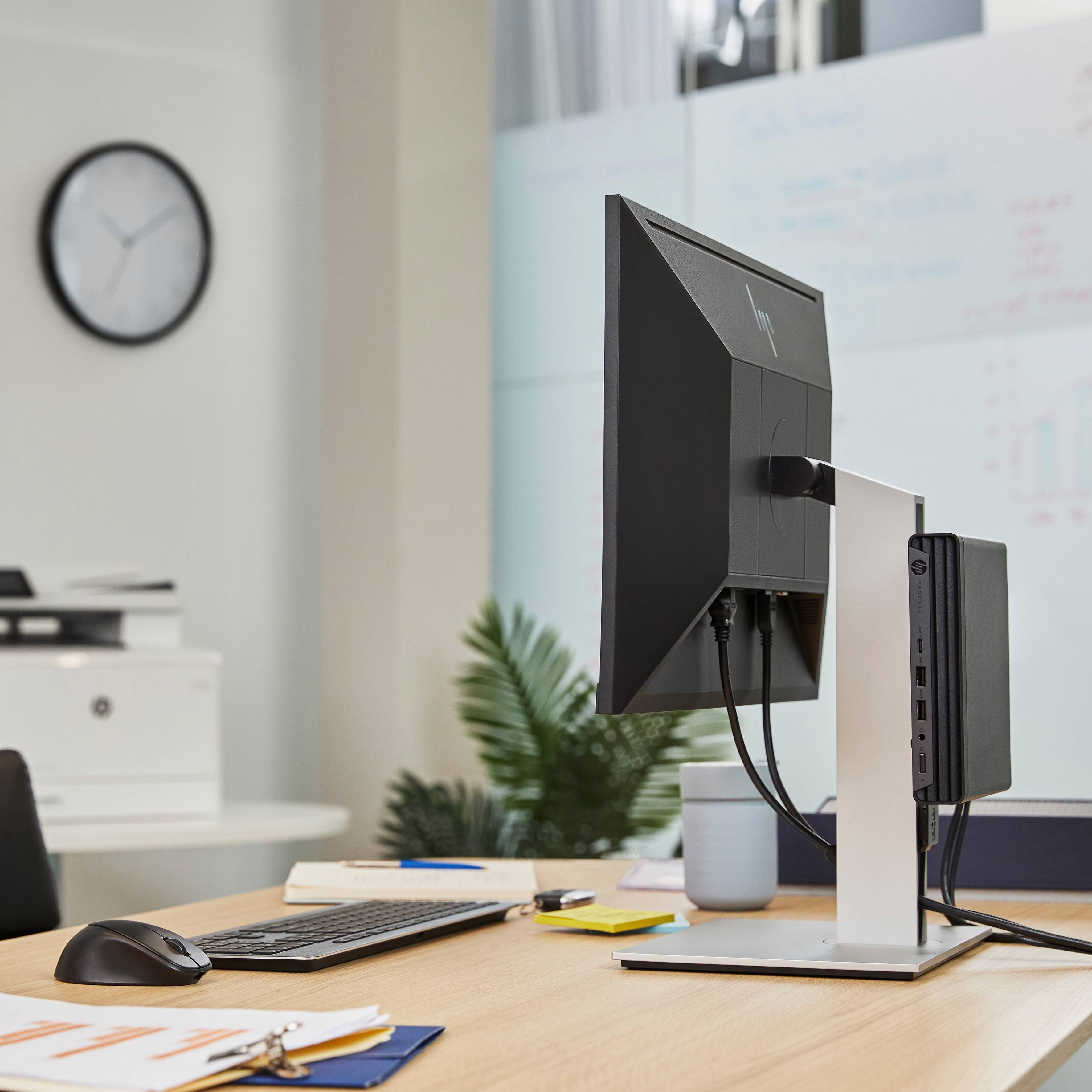Büroarbeitsplatz mit Computer, Tastatur, Notizen und Kaffeetasse. Im Hintergrund eine Wanduhr und ein Whiteboard mit Grafiken.