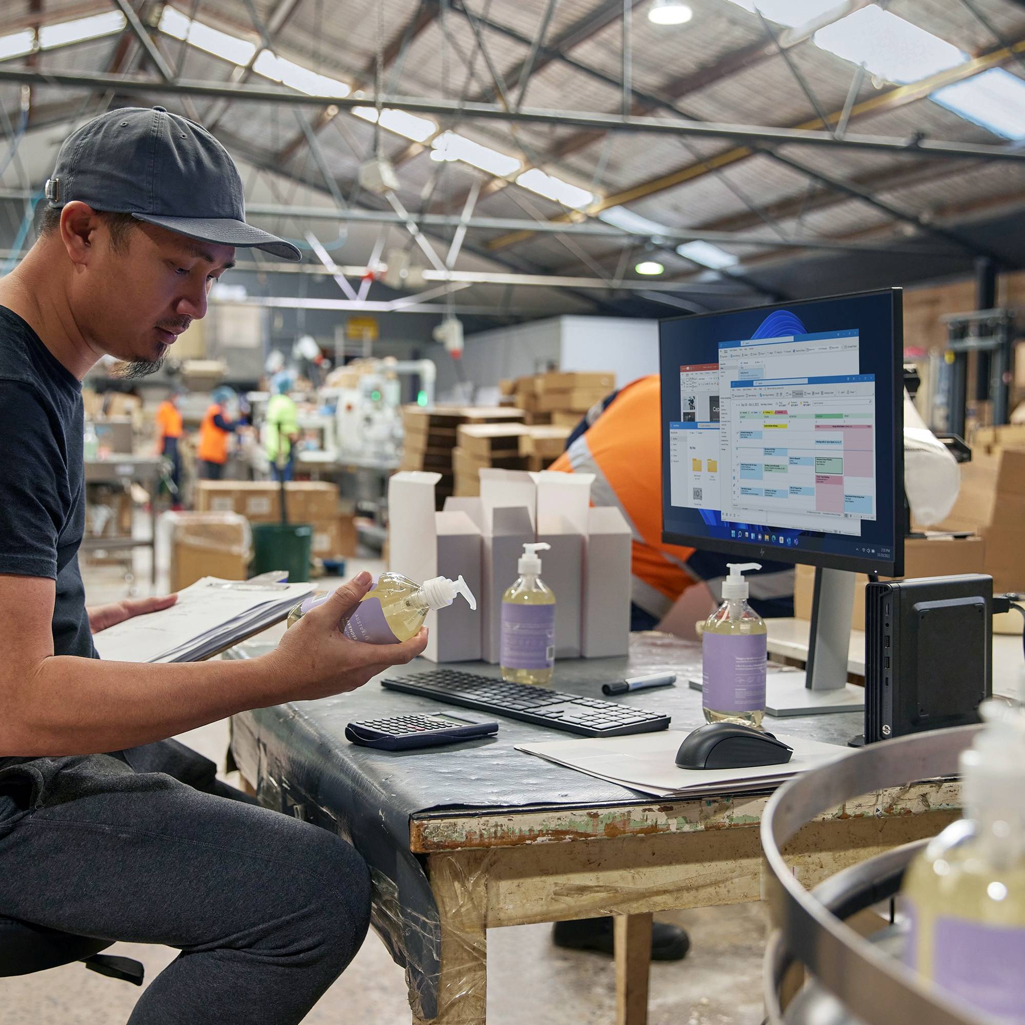 Ein Mann in Arbeitskleidung sitzt an einem Schreibtisch in einer Fabrikhalle, überprüft Kartons und Daten auf einem Computerbildschirm. Im Hintergrund sind weitere Arbeiter tätig.