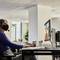 Eine Person mit Headset sitzt an einem Schreibtisch, schaut auf einen Computerbildschirm mit einem Videogespräch, neben ihr ein Getränk.
