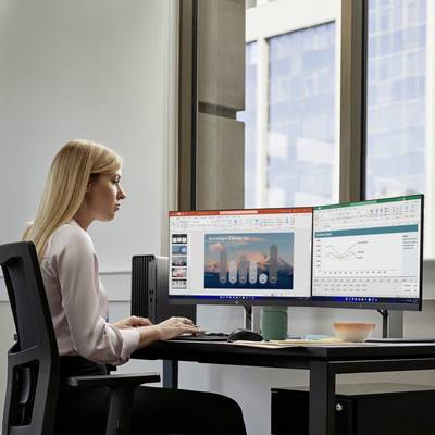 Eine Frau sitzt an einem Schreibtisch und arbeitet an zwei Computerbildschirmen. Auf den Monitoren sind Diagramme und Präsentationen zu sehen.