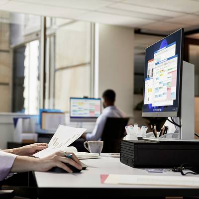 Büroumgebung mit zwei Mitarbeitern an Computern, Bildschirme zeigen Kalender- und Planungssoftware. Gerichtete Hände auf Dokumenten.