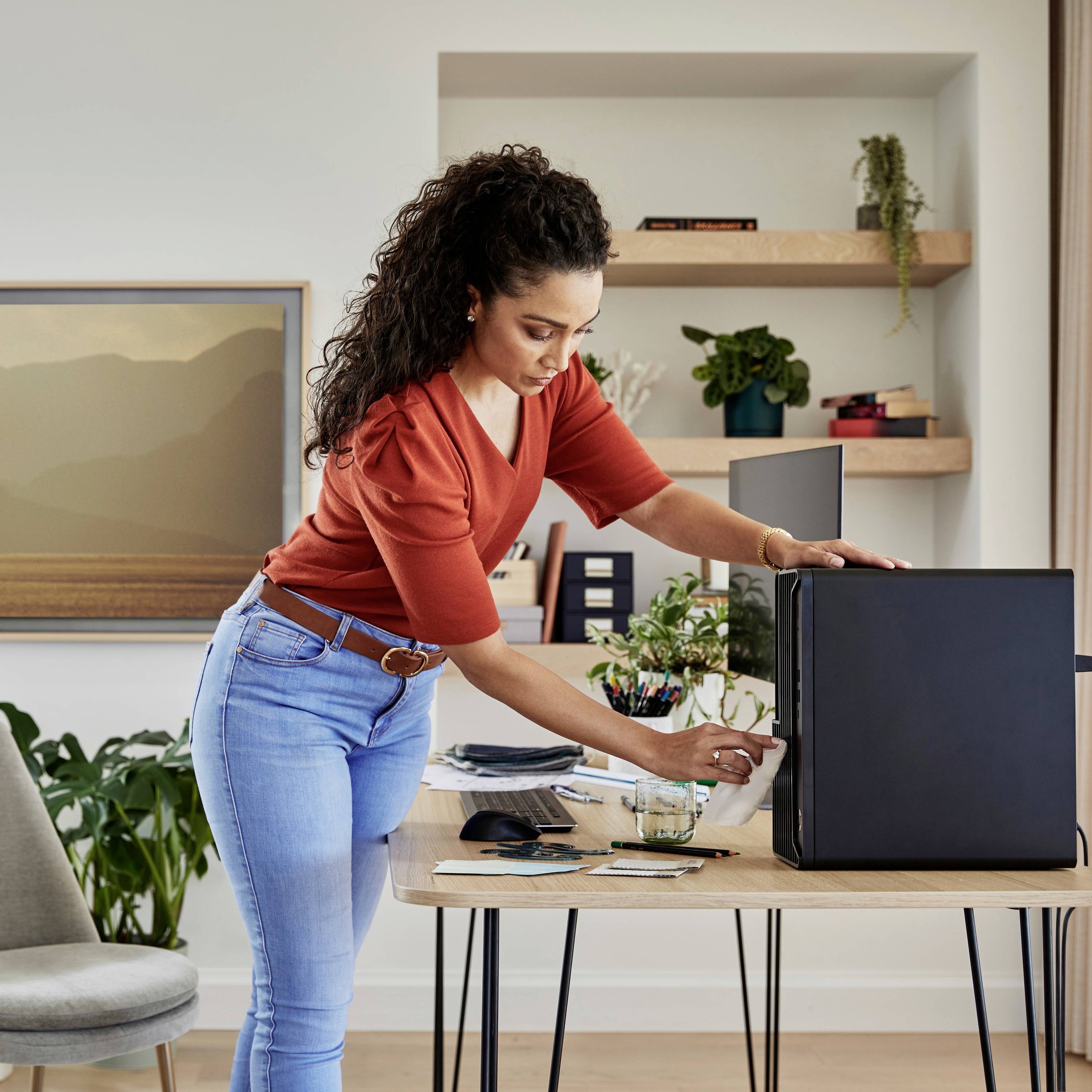 Eine Frau in einem roten Pullover wischt staub von einem schwarzen Computer in einem modernen Büro mit Pflanzen und Kunstwerken.