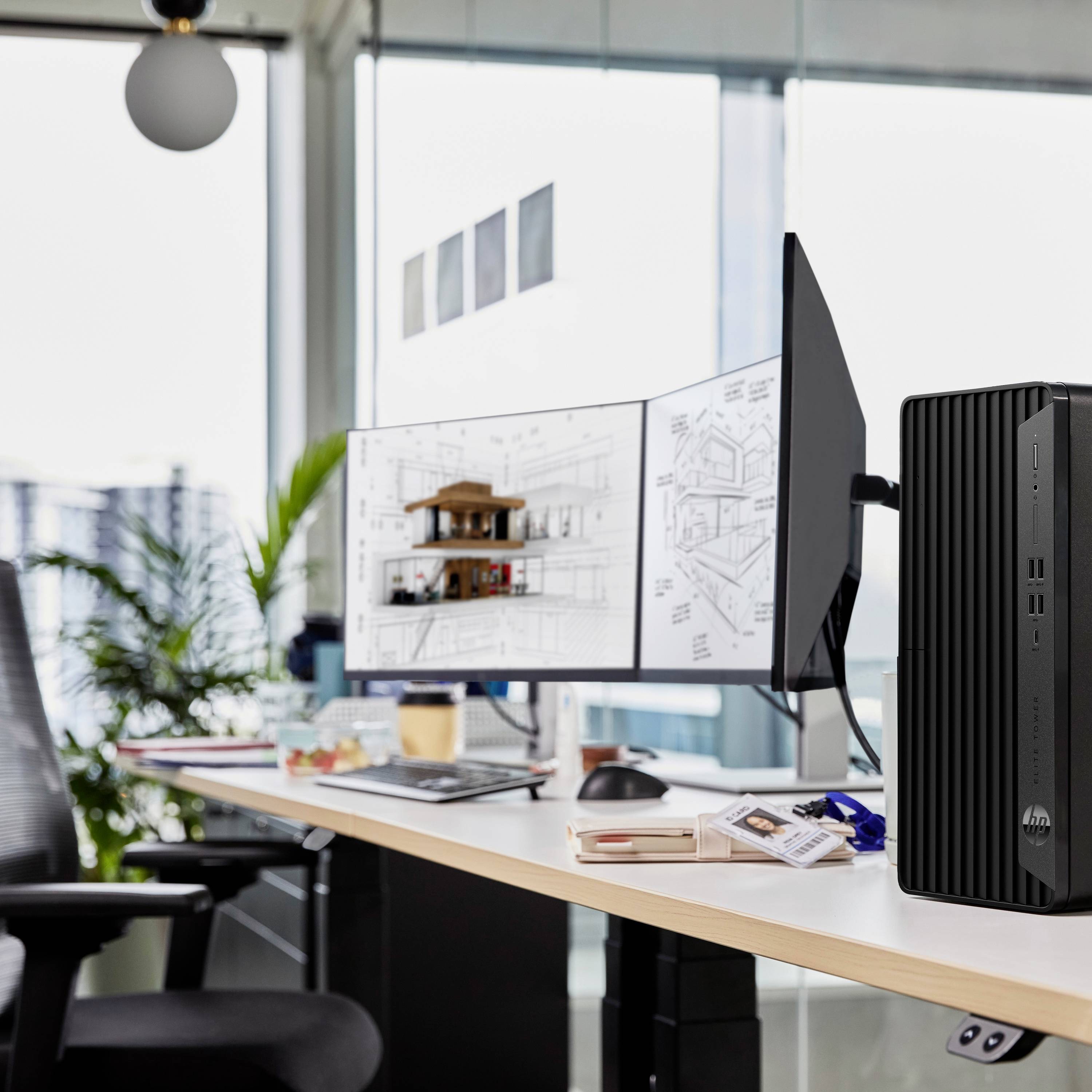 Ein Schreibtisch mit Monitor zeigt Architekturentwürfe. Ein Computer steht daneben. Im Hintergrund Pflanzen und ein Fenster mit Stadtblick.