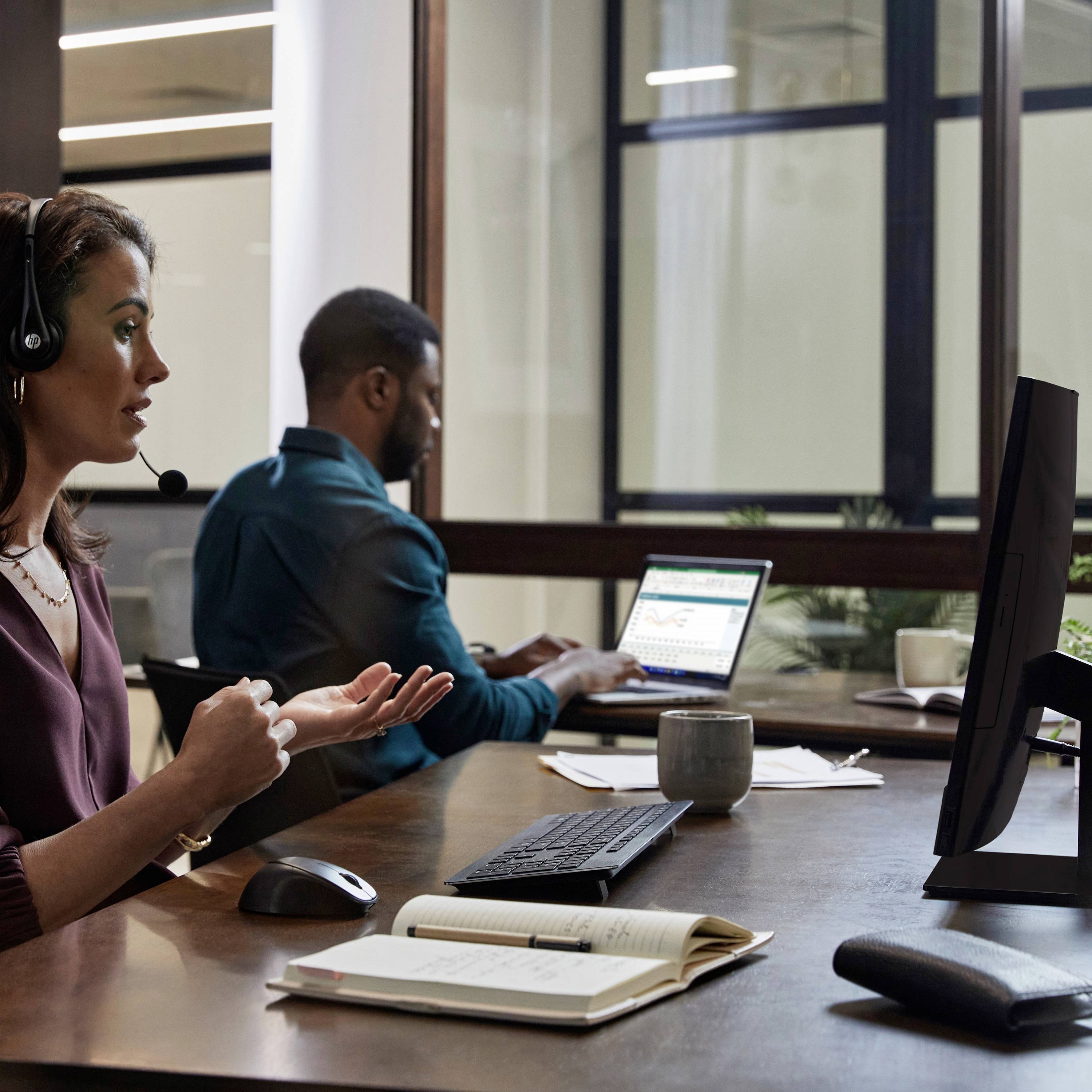 Eine Frau mit Headset spricht gestikulierend, während ein Mann im Hintergrund an einem Laptop arbeitet. Sie befinden sich in einem modernen Büro.
