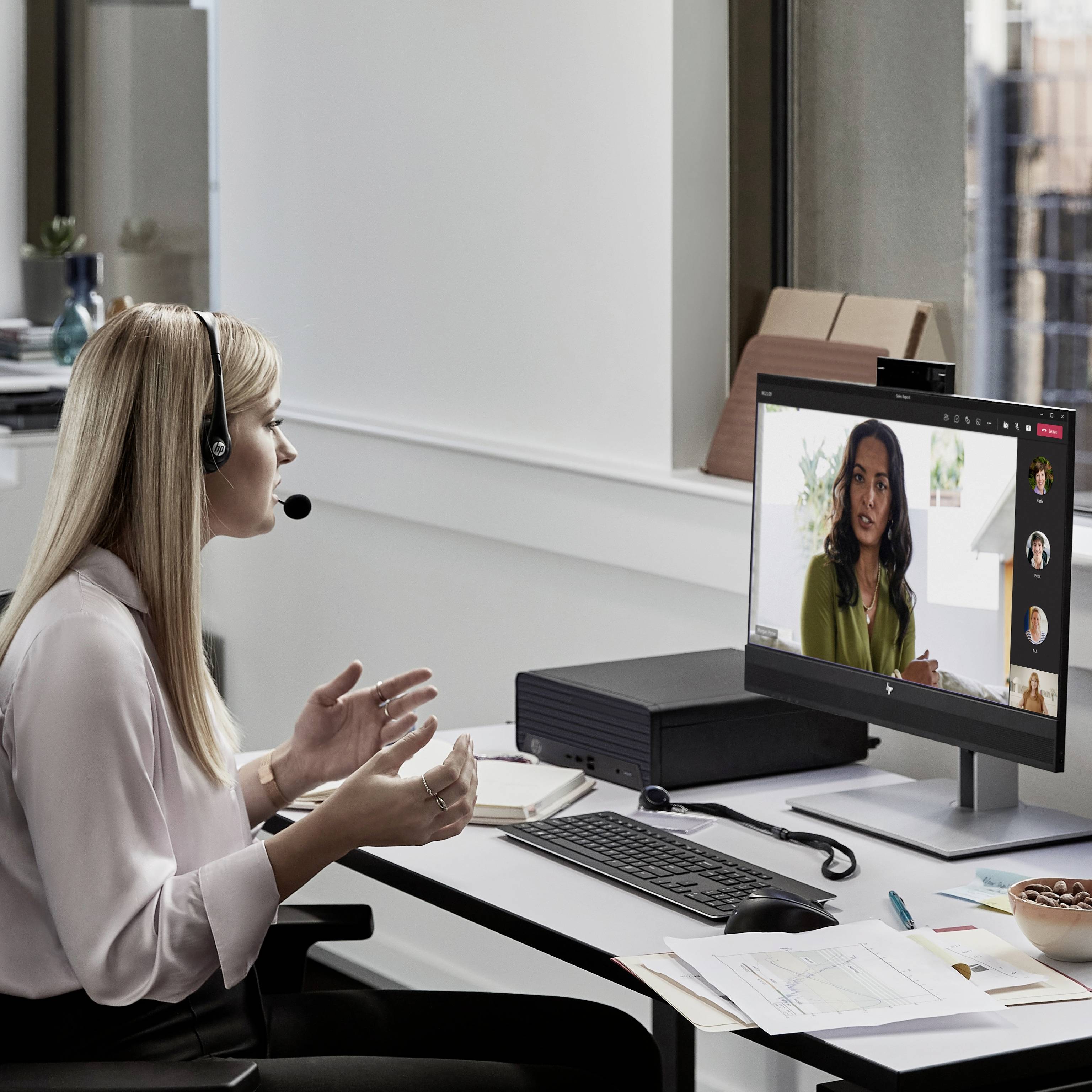 Eine Frau mit Headset sitzt an einem Schreibtisch und nimmt an einer Videokonferenz auf einem Monitor teil, der mehrere Teilnehmer zeigt.