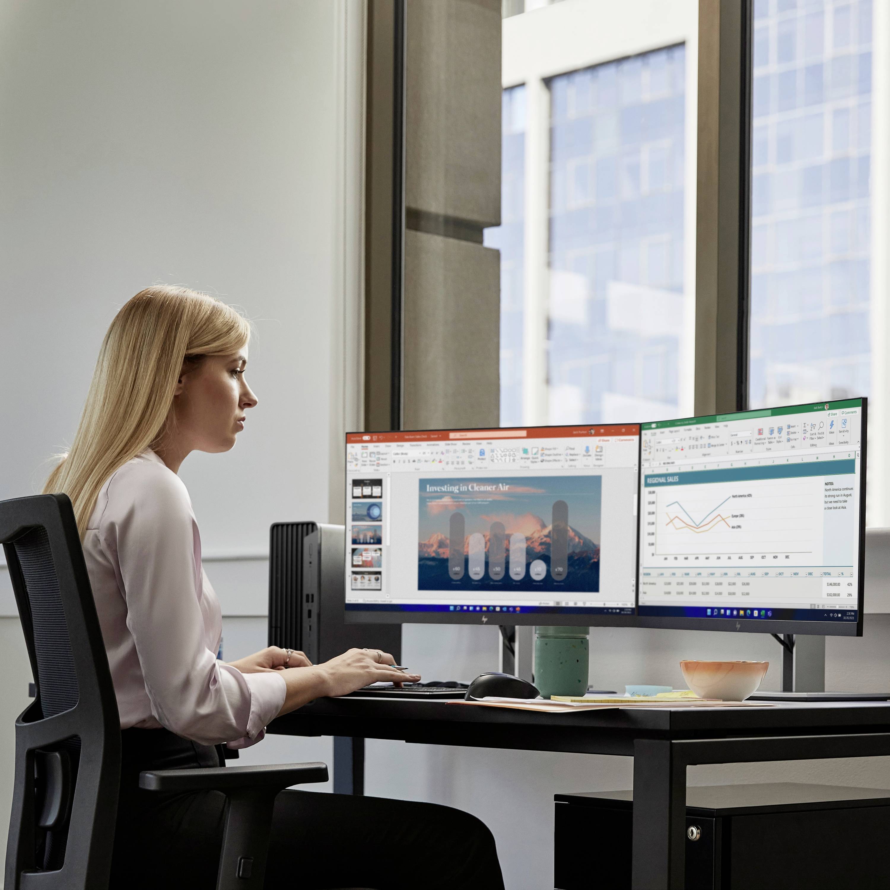 Eine Frau sitzt an einem Schreibtisch und arbeitet an einem Computer mit zwei Bildschirmen. Grafiken und Dokumente sind zu sehen.