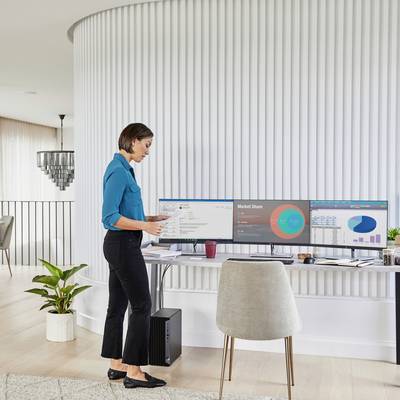 Eine Frau in einem blauen Hemd steht an einem Schreibtisch, betrachtet zwei Computermonitore mit Marktanteildiagrammen und Daten.