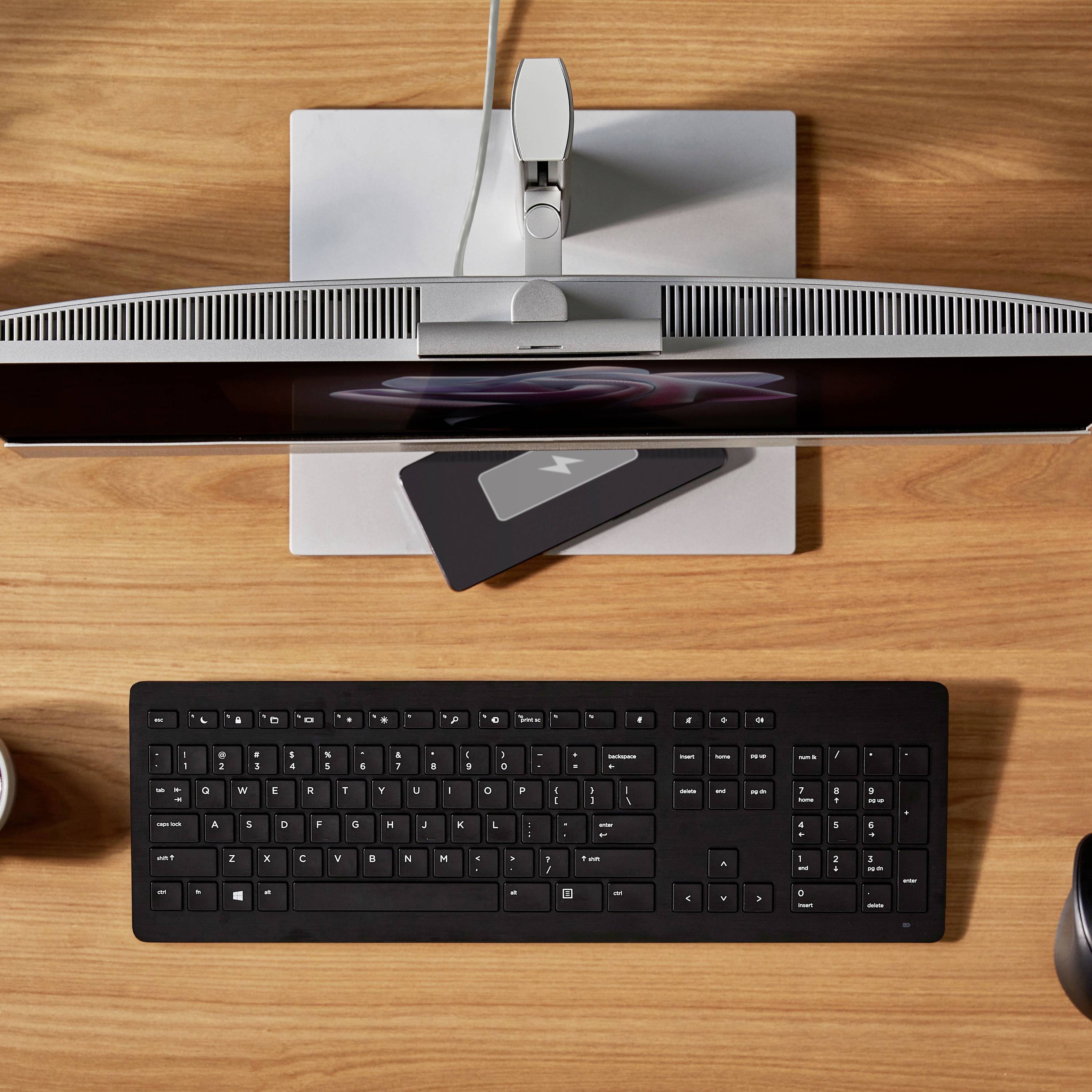 Ein moderner Arbeitsplatz mit Monitor, Tastatur und Ladegerät für Mobilgeräte auf einem Holztisch; Tasse an der Seite.