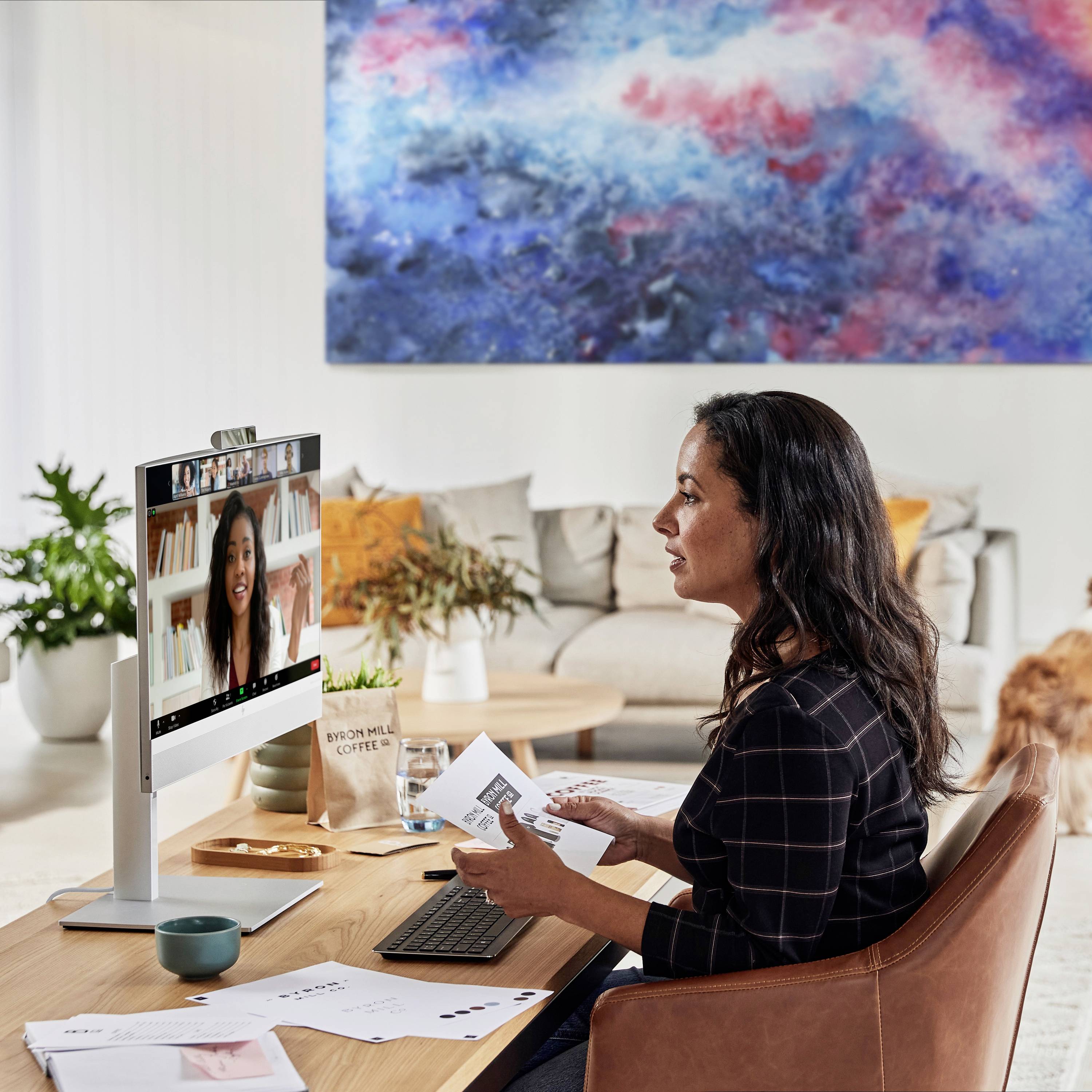 Eine Frau sitzt an einem Schreibtisch im Homeoffice und führt ein Videogespräch. Im Hintergrund sind ein Sofa und ein großes Kunstbild zu sehen.