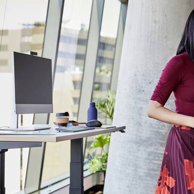 Die Person steht an einem Steharbeitsplatz mit Laptop, Kaffeetasse und Wasserflaschen. Hintergrund: moderne Bürofenster.