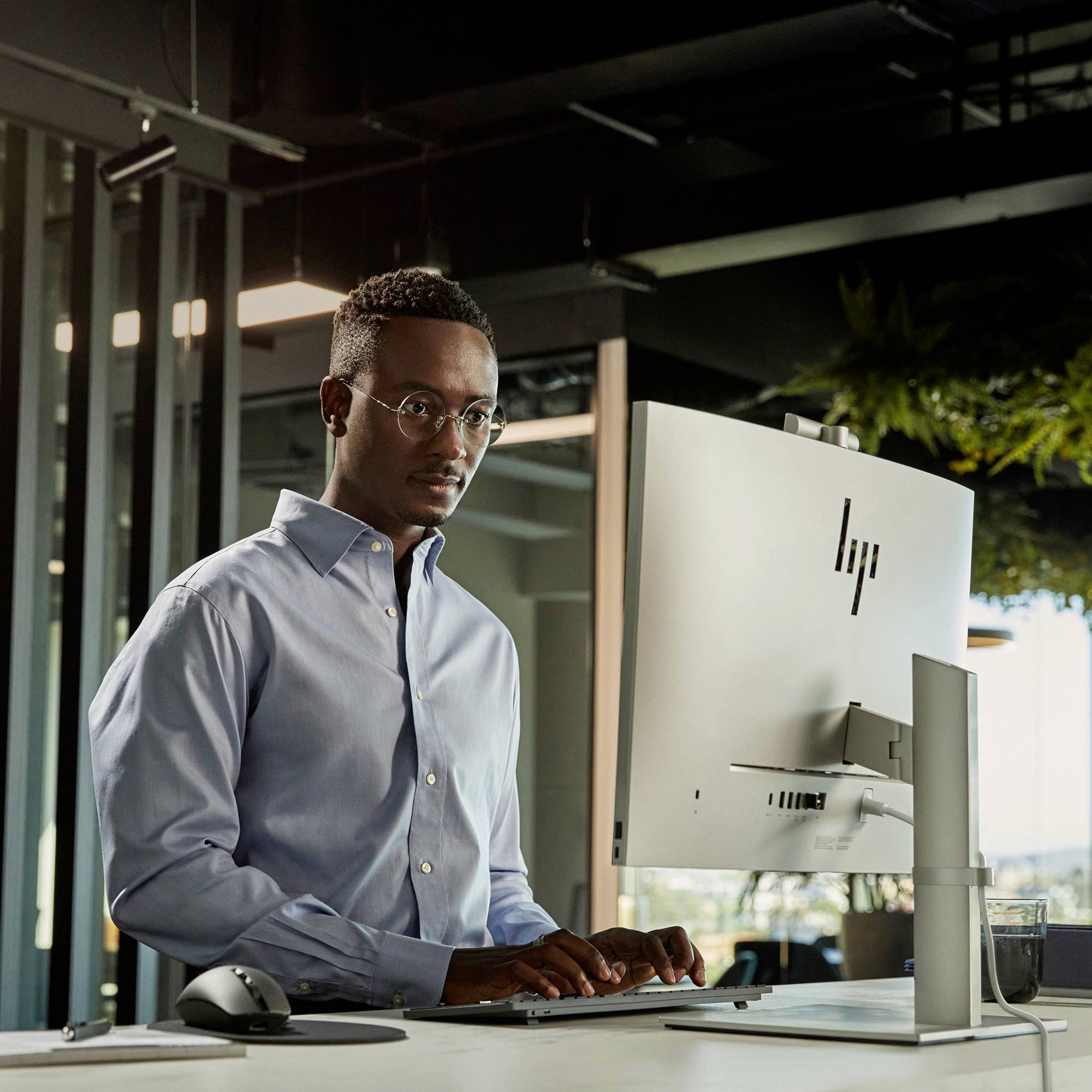 Eine Person arbeitet an einem Schreibtisch mit einem Computer in einem modernen Büro. Sie trägt ein hellblaues Hemd und schaut auf den Bildschirm.