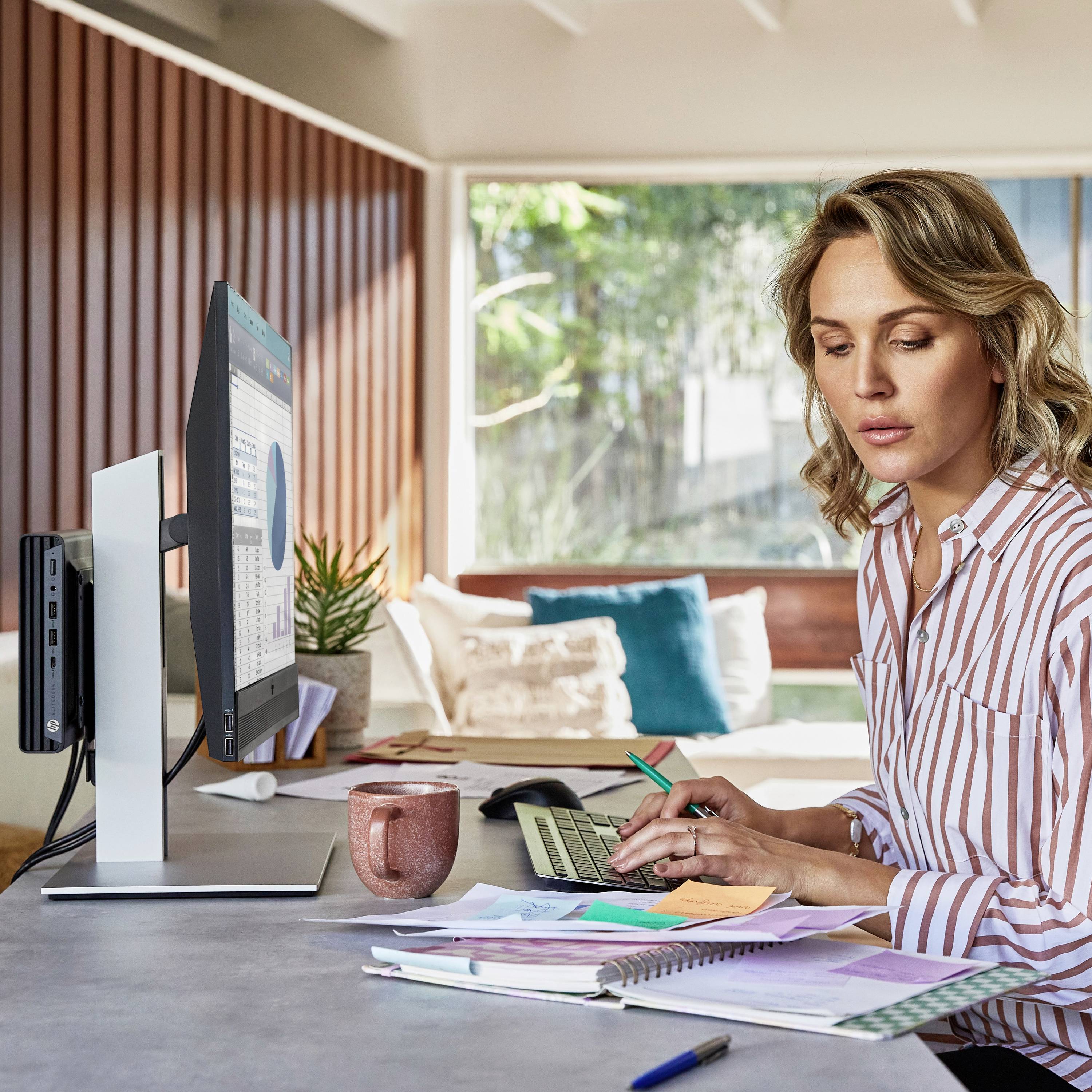 Eine Frau sitzt an einem Schreibtisch mit Computer und schreibt in einem Notizbuch. Im Hintergrund ist ein Sofa mit Kissen zu sehen.