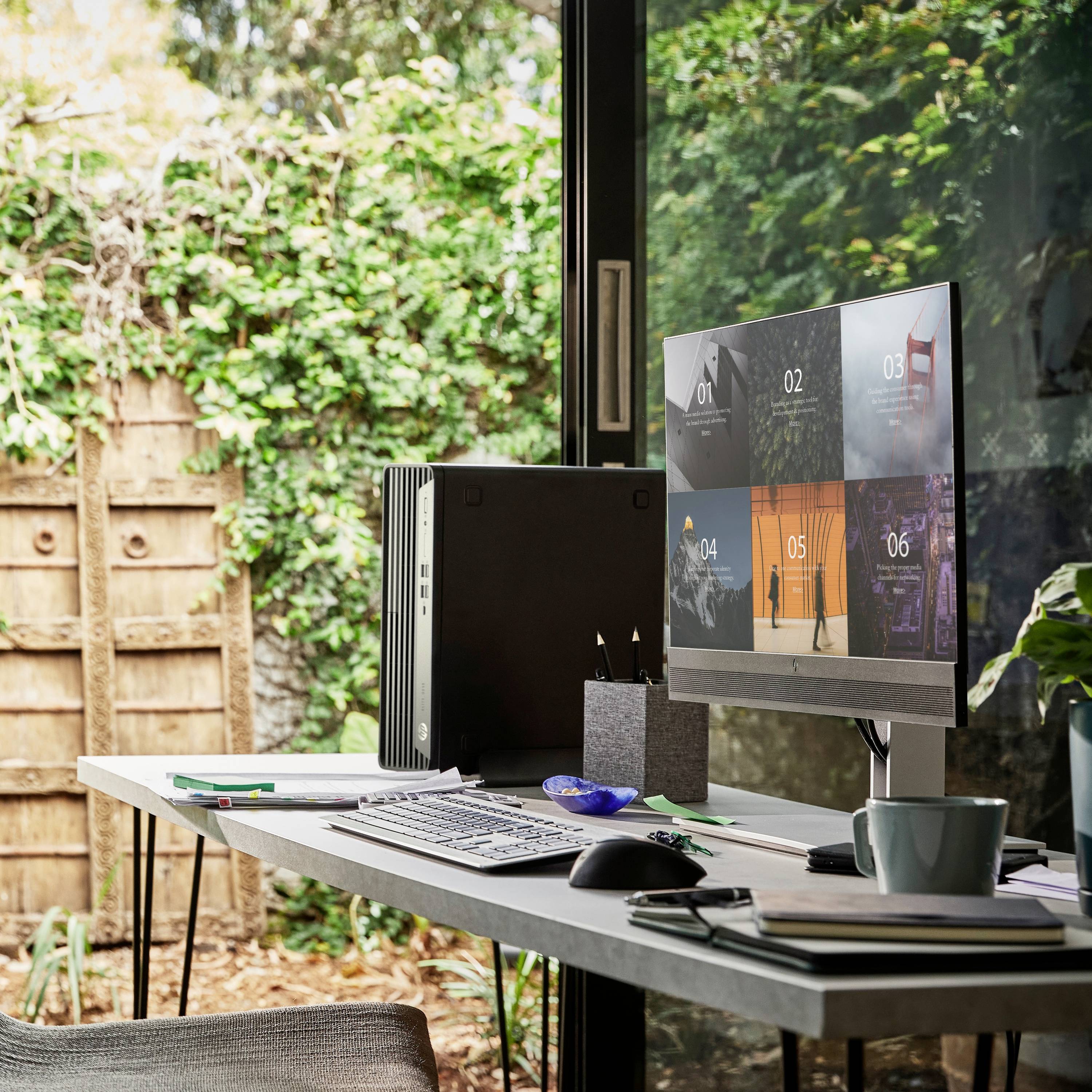 Moderner Arbeitsplatz mit Blick auf einen Garten. Computerbildschirm zeigt mehrere Bildschirme. Auf dem Tisch liegen Notizbücher und Stifte.