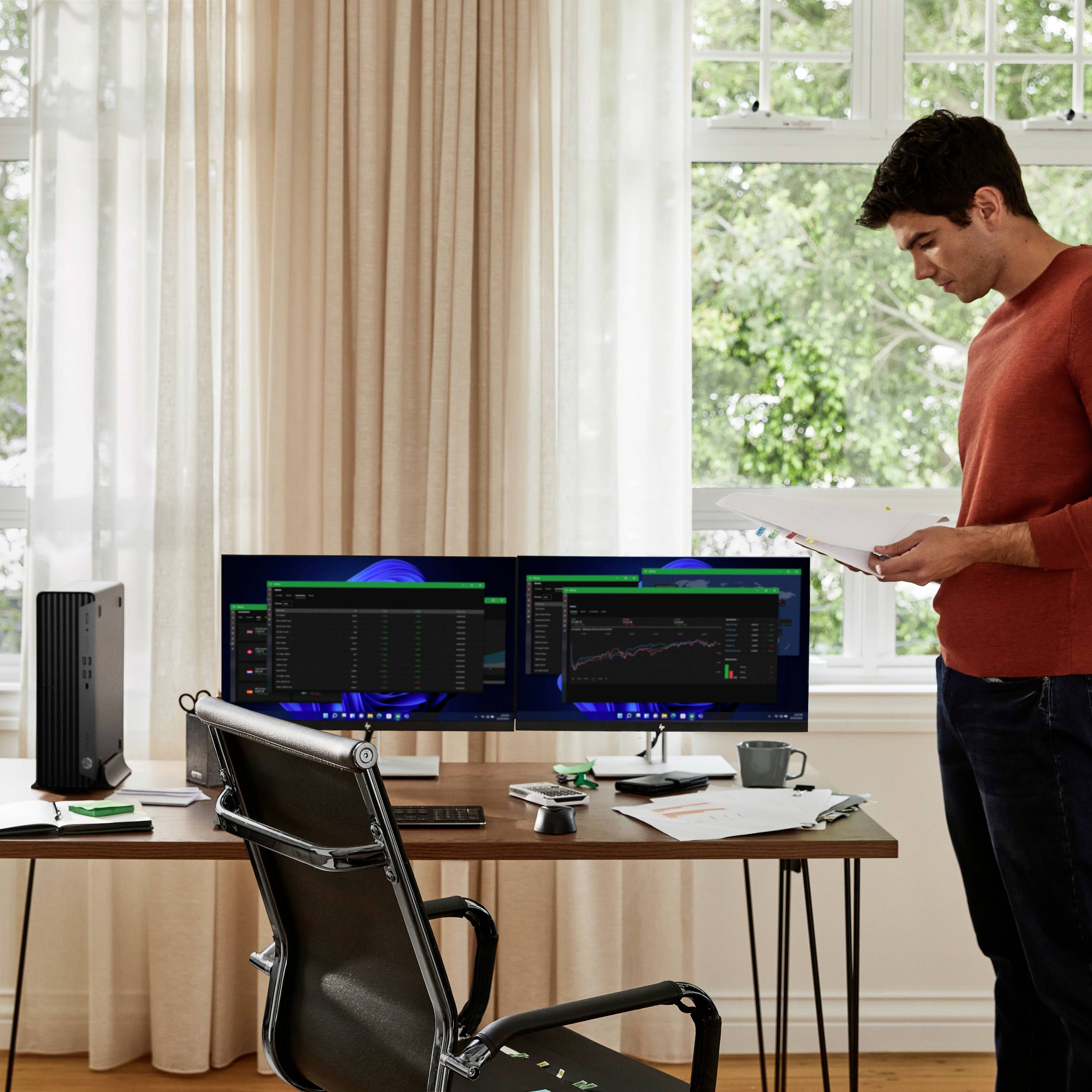 Ein Mann in einem roten Pullover betrachtet Dokumente vor einem Schreibtisch mit zwei Monitoren und einem Computer, im Hintergrund Fenster.