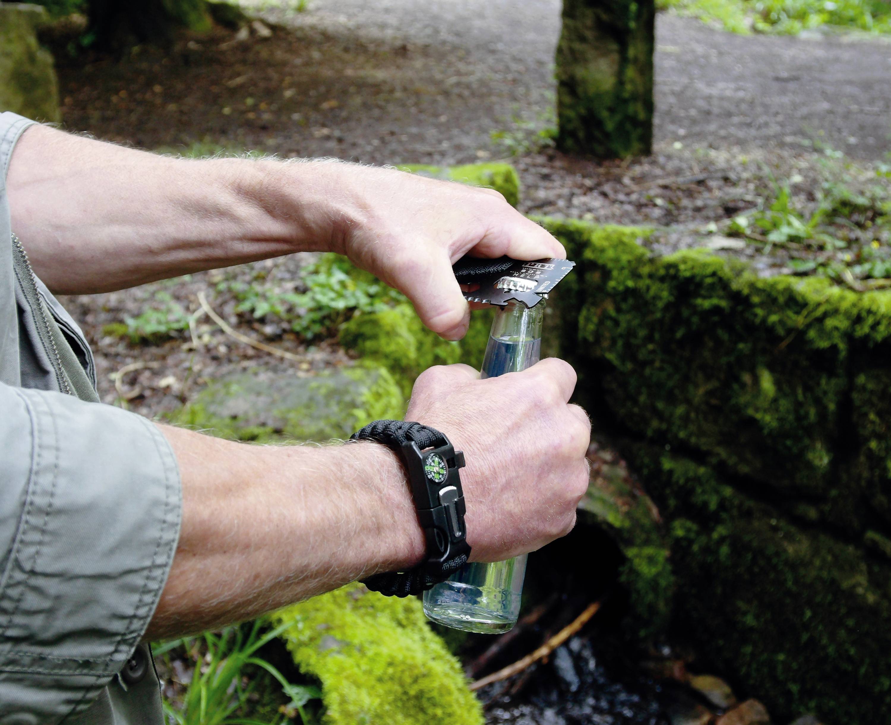 Eine Person öffnet eine Glasflasche mit einem schwarzen Flaschenöffner im Freien, umgeben von grüner Vegetation und einem steinernen Hintergrund.