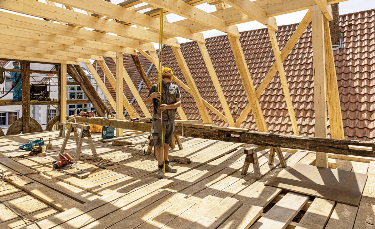 Ein Zimmermann steht auf einer Baustelle und arbeitet an einem Dachstuhl aus Holz. Im Hintergrund sind Ziegeldächer zu sehen.