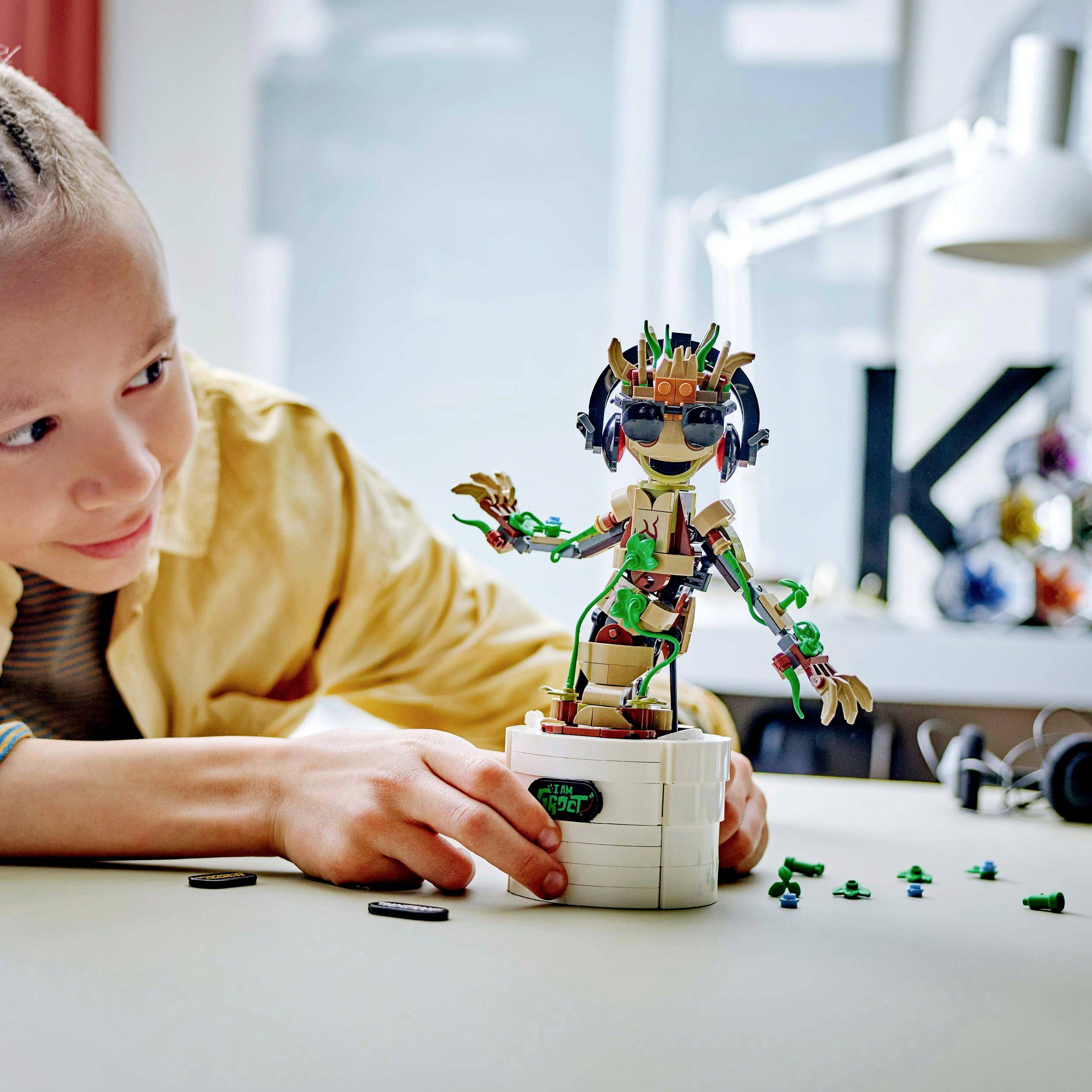 Ein Kind betrachtet lächelnd einen bunten Spielzeugroboter auf einem Tisch. Der Roboter hat Sonnenbrille und Kopfhörer.