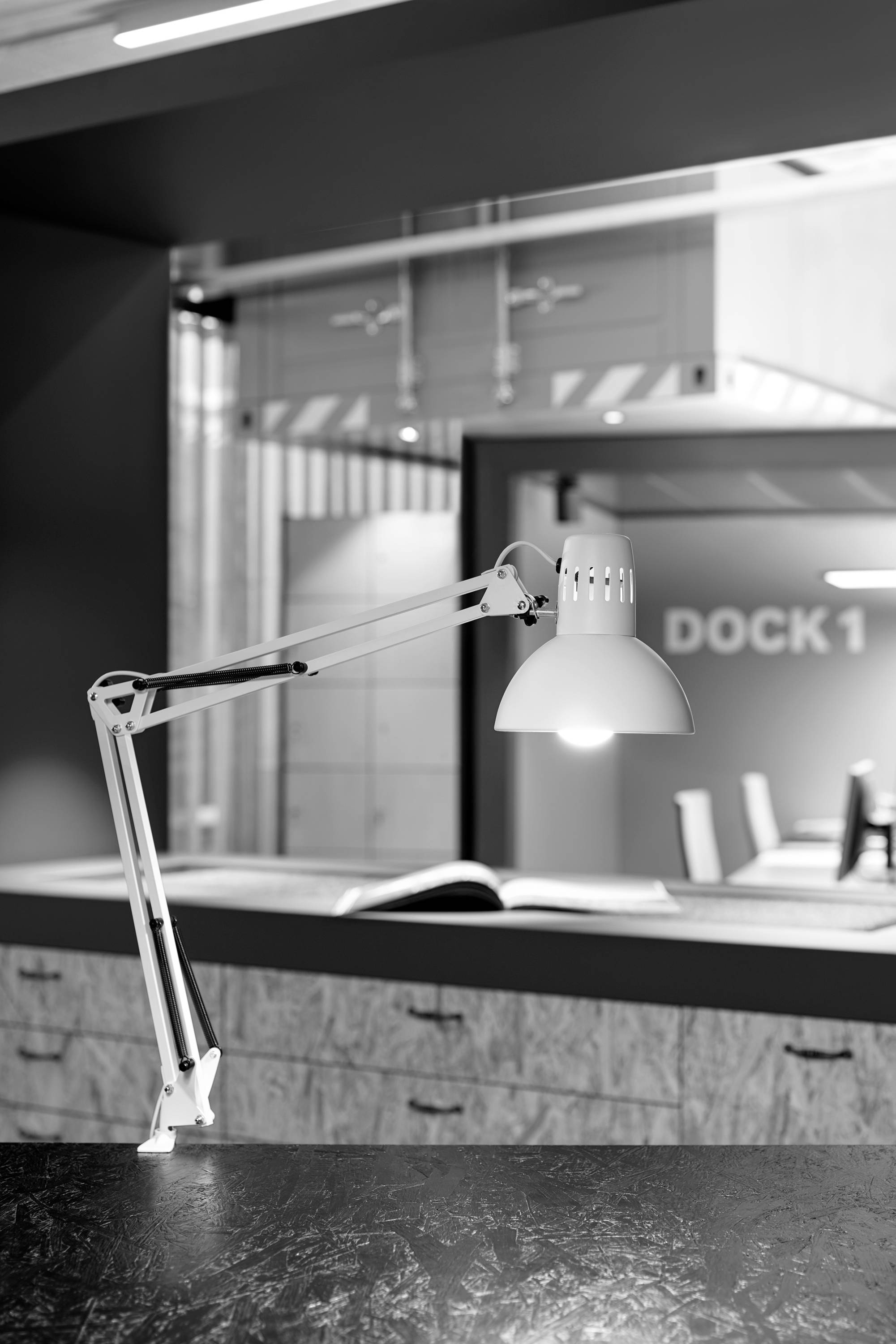 Schwarz-weiß Bild zeigt eine moderne Schreibtischlampe auf einem Tisch im Büro. Im Hintergrund sind Regale und der Schriftzug 'DOCK 1' sichtbar.