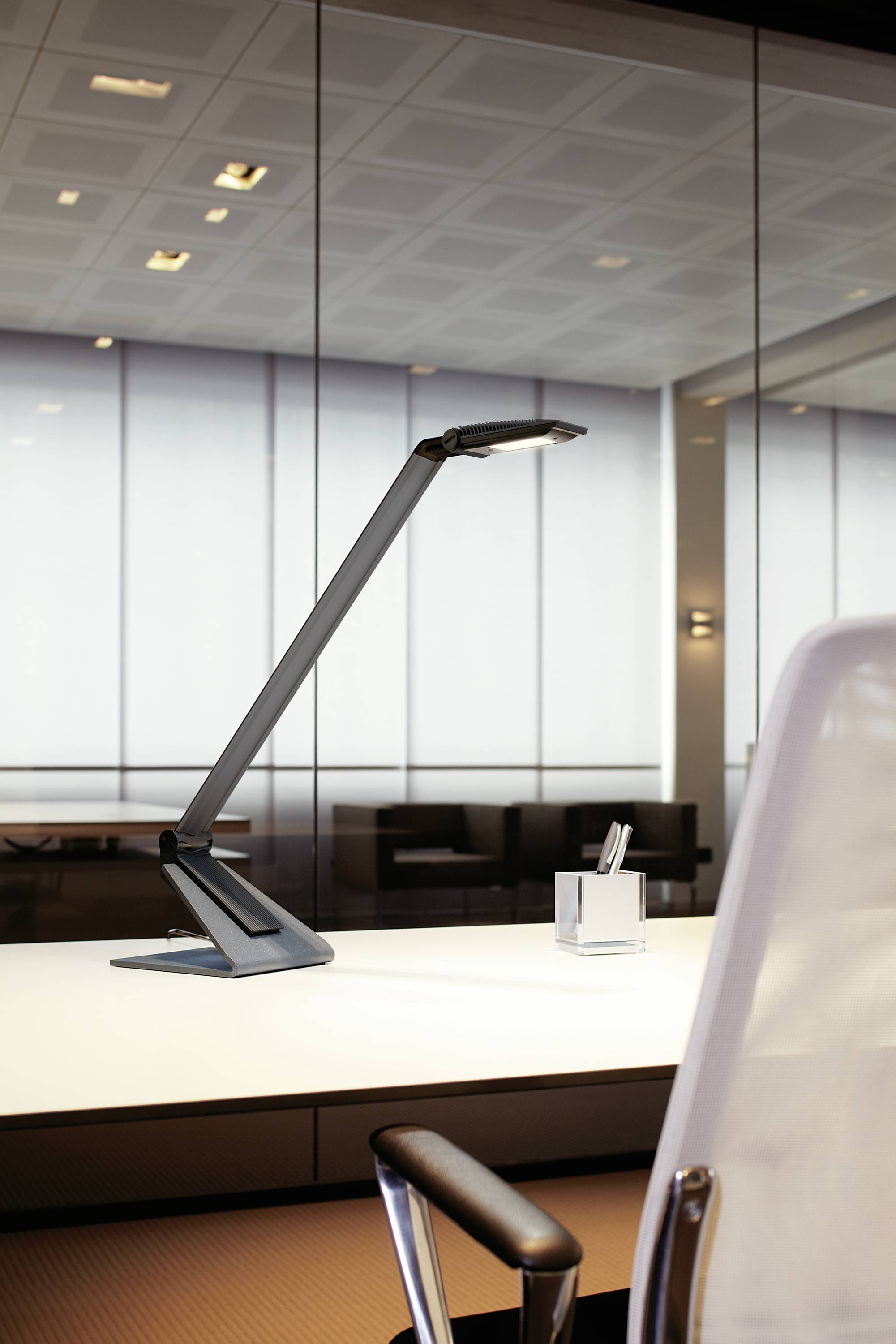 Büroszene mit modernem Schreibtisch, einer eleganten Tischlampe und einem Stuhl. Hintergrund aus Glaswänden und gedämpftem Licht.