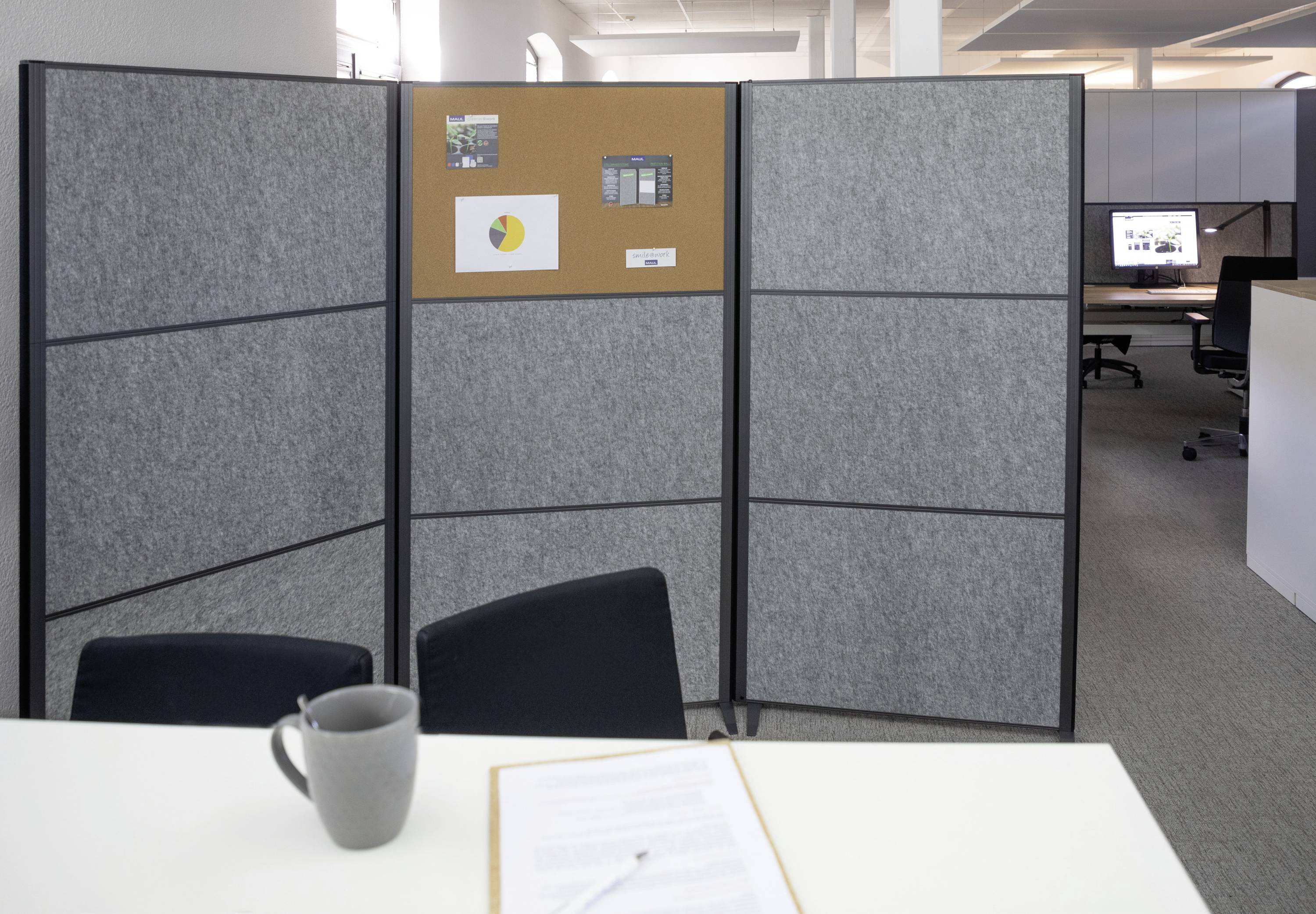 Büroraum mit grauem Stellwandteiler, der zwei Arbeitsbereiche trennt. Darauf Korktafel mit Notizen. Im Vordergrund ein Schreibtisch mit Tasse und Dokument.