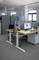 Ein modernes Büro mit höhenverstellbaren Schreibtischen, Bildschirm und Laptops. Sichtschutzwände trennen die Arbeitsplätze.