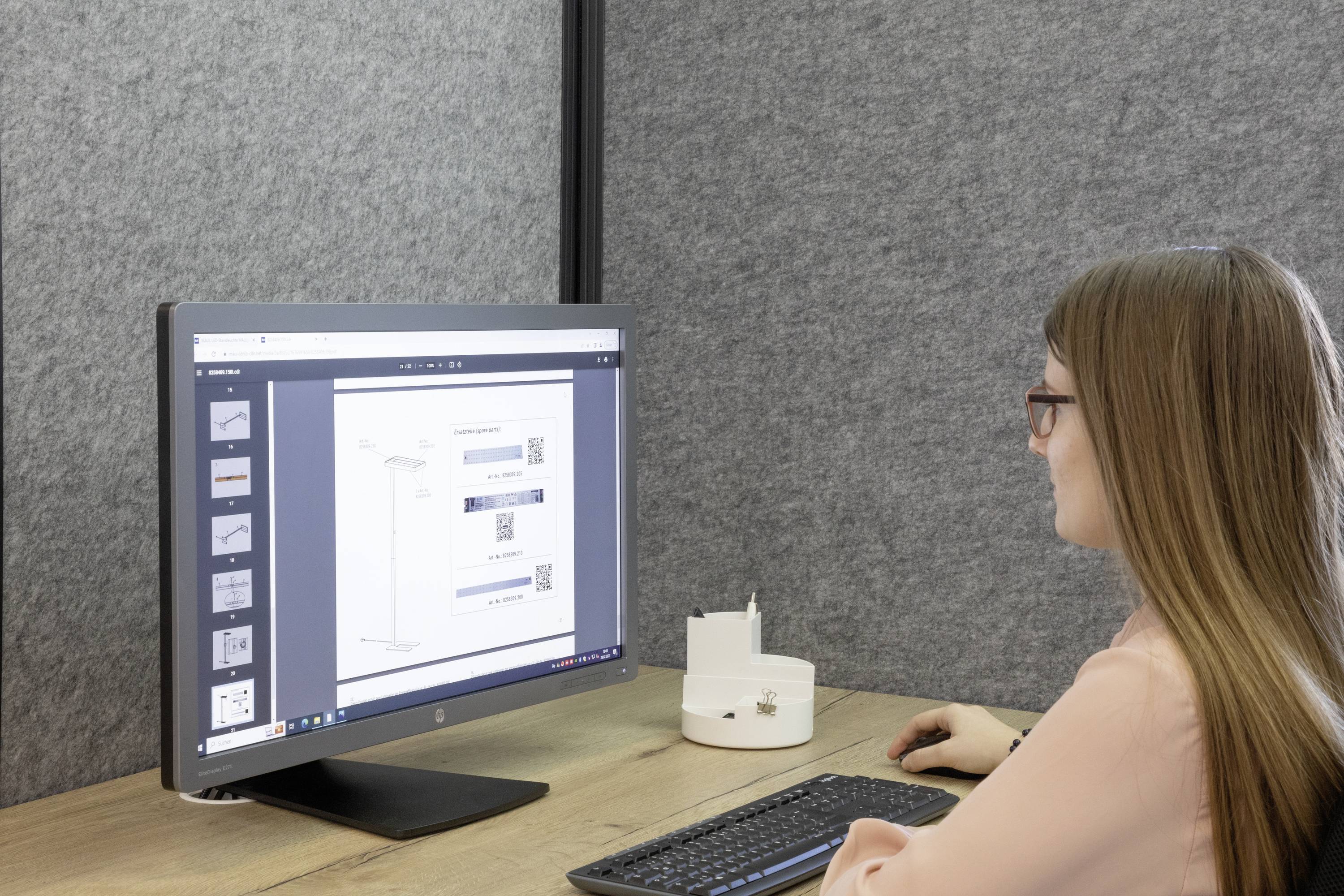Eine Frau mit langen Haaren arbeitet an einem Computer, der ein Dokument anzeigt. Der Raum ist mit grauem Stoff verkleidet.
