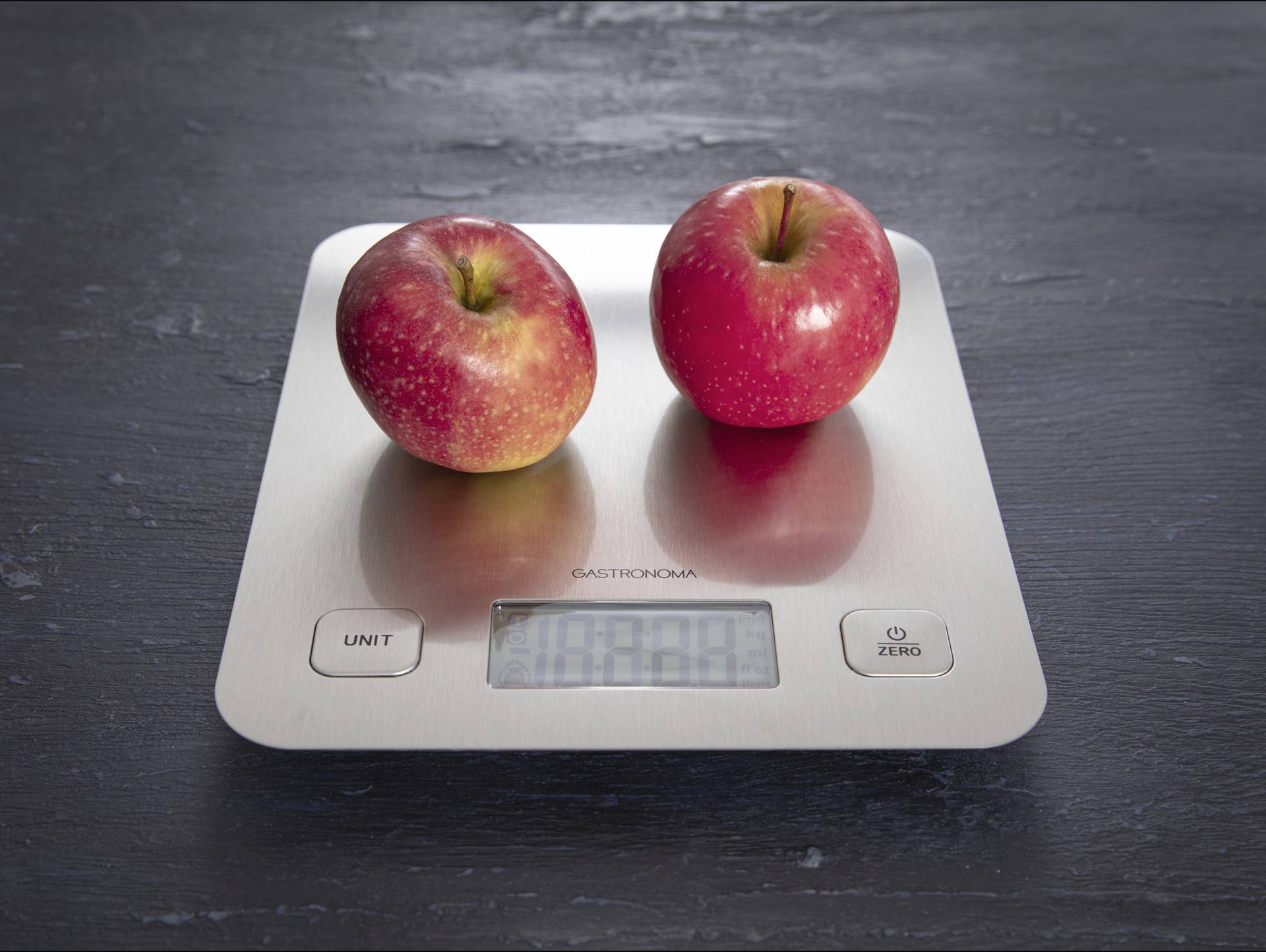 Zwei rote Äpfel liegen auf einer digitalen Küchenwaage auf einer dunklen Oberfläche. Die Waage zeigt ein Gewicht an.