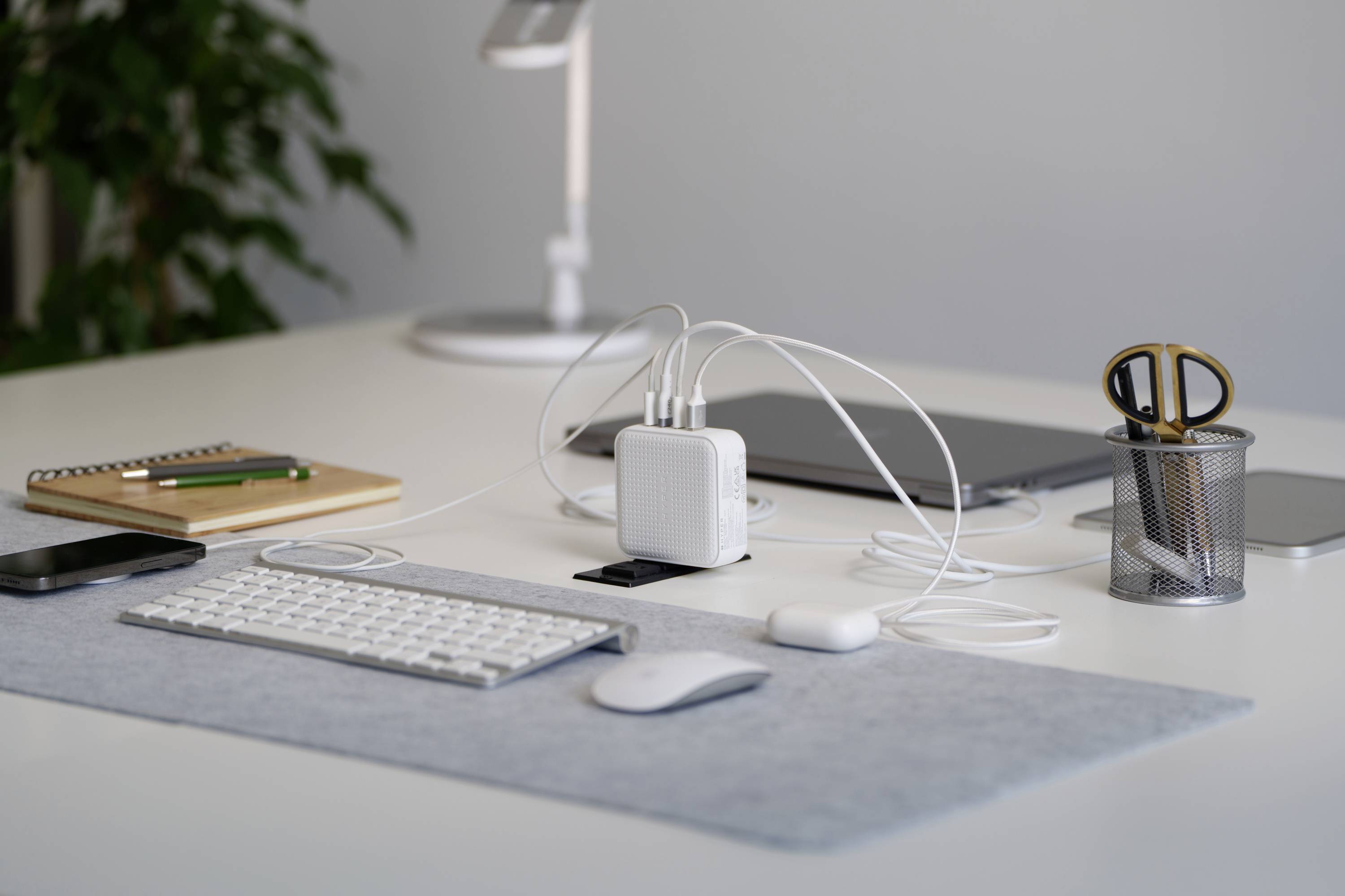 Ein ordentlich aufgeräumter Schreibtisch mit einer Tastatur, Maus, Ladegerät mit mehreren Kabeln, Notizbuch, Pflanzen und Laptop im Hintergrund.