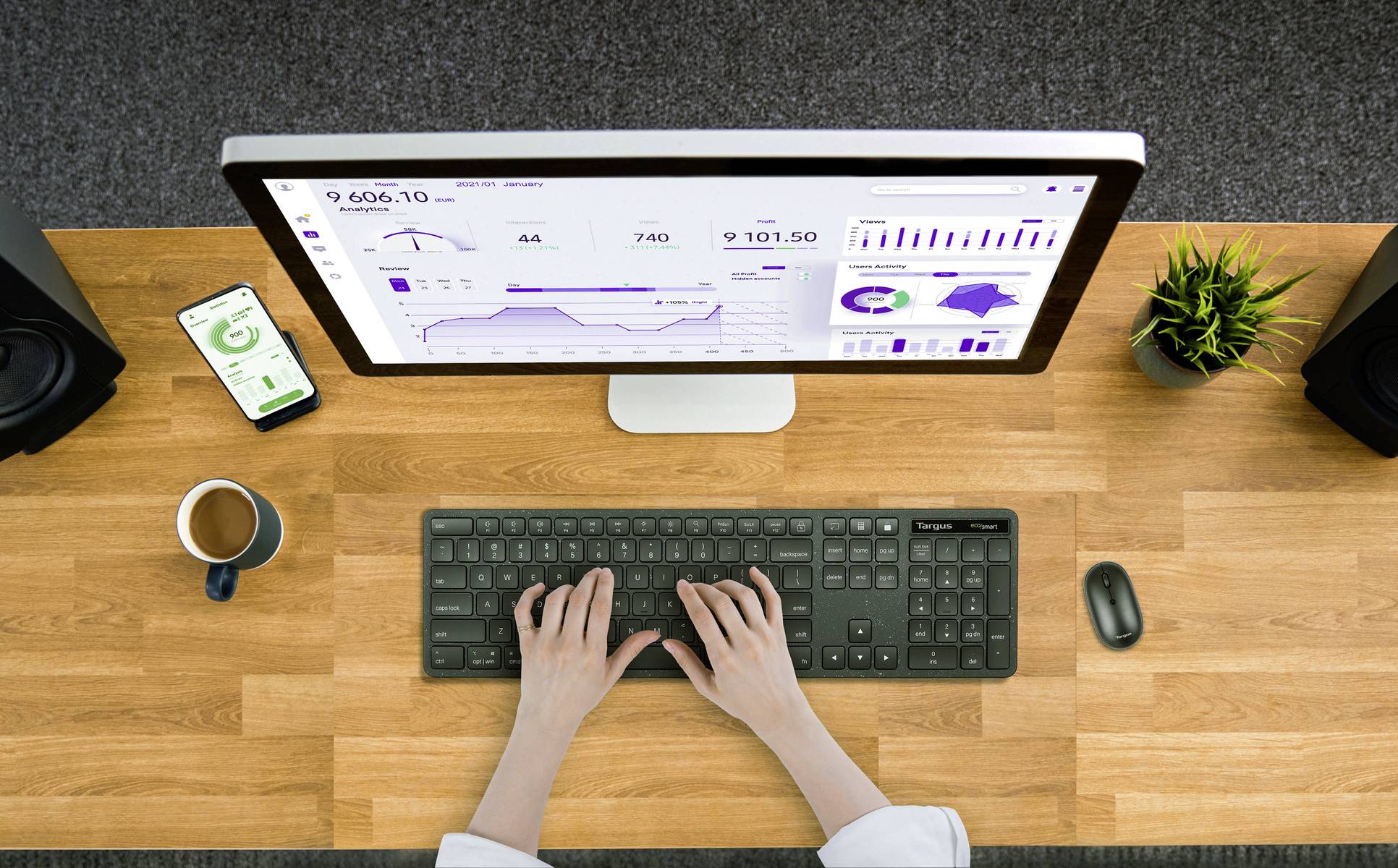 Eine Person tippt auf einer Tastatur vor einem Monitor mit Datencharts. Ein Smartphone, eine Tasse Kaffee und Pflanzen befinden sich auf dem Schreibtisch.