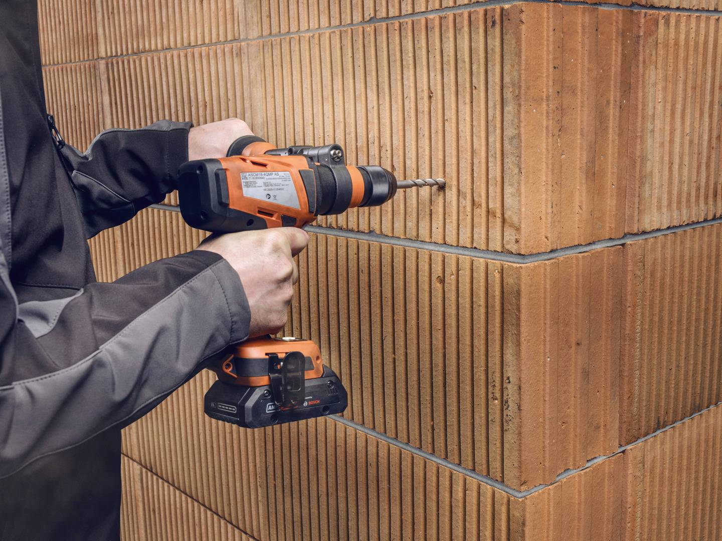 Eine Person verwendet eine orange-braune Bohrmaschine, um in eine Ziegelwand zu bohren. Die Wand besteht aus strukturierten Backsteinen.