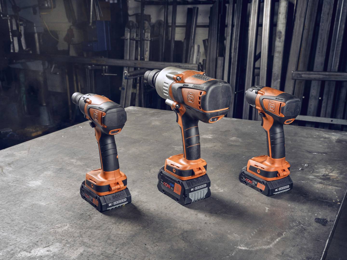 Drei orange-schwarze Akkuschrauber in unterschiedlichen Größen stehen auf einem Metalltisch in einer industriellen Umgebung.