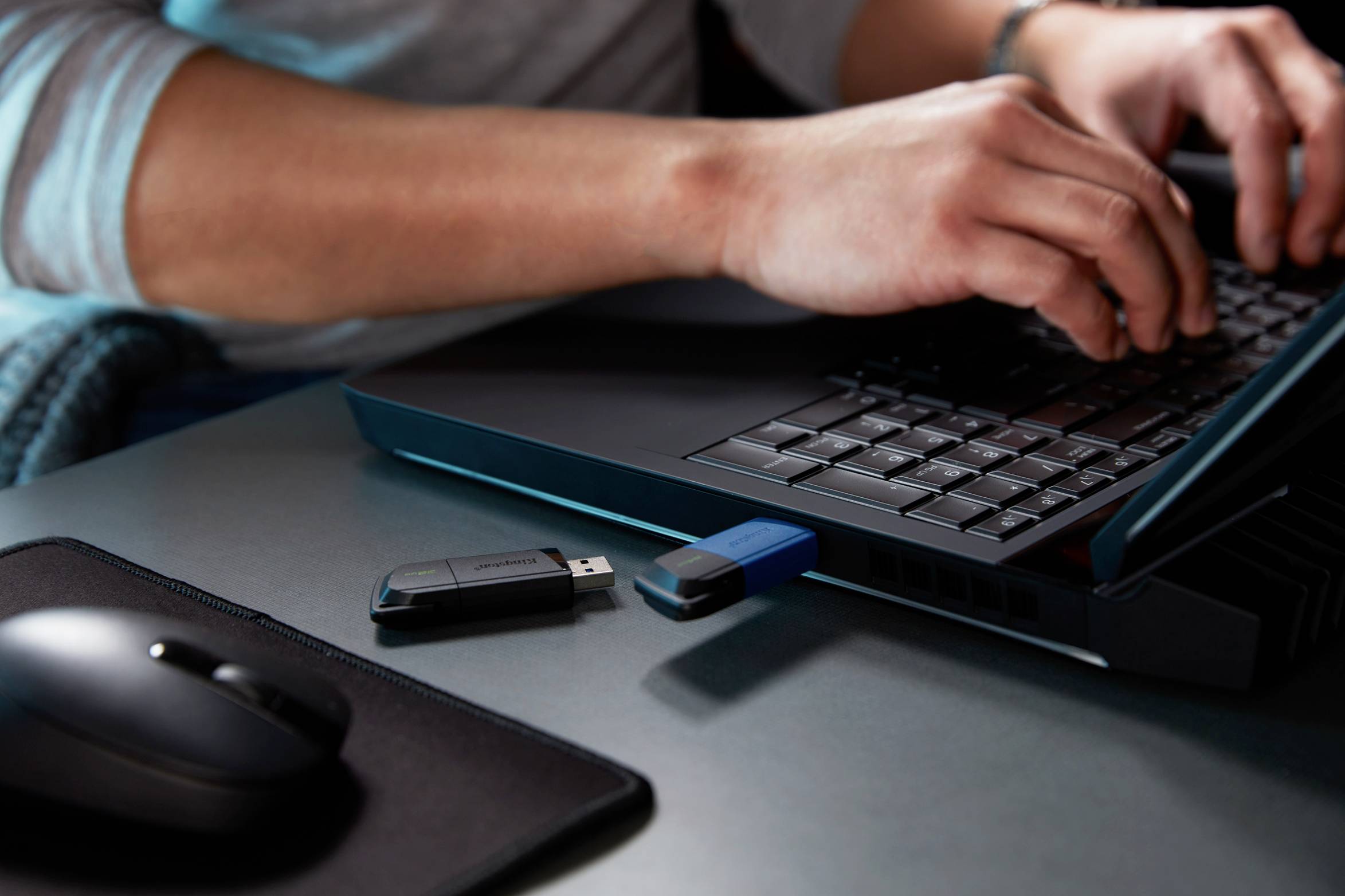 Person tippt auf einem Laptop. Zwei USB-Sticks sind in den Laptop eingesteckt. Rechts liegt eine Computermaus auf einem Mauspad.
