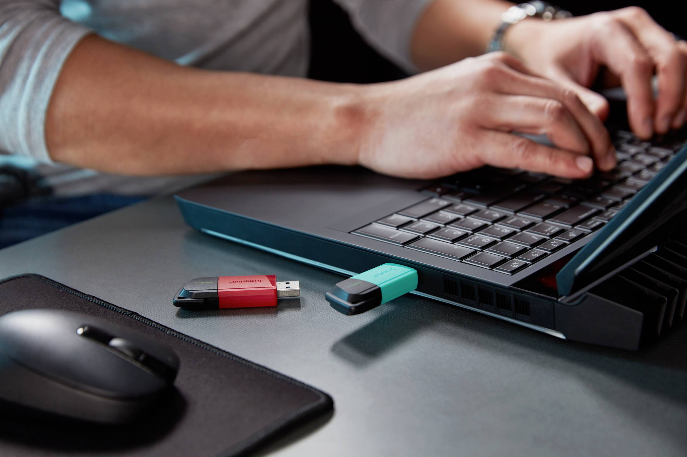 Person tippt auf Laptop, zwei USB-Sticks liegen daneben auf dem Tisch, einer ist eingesteckt.