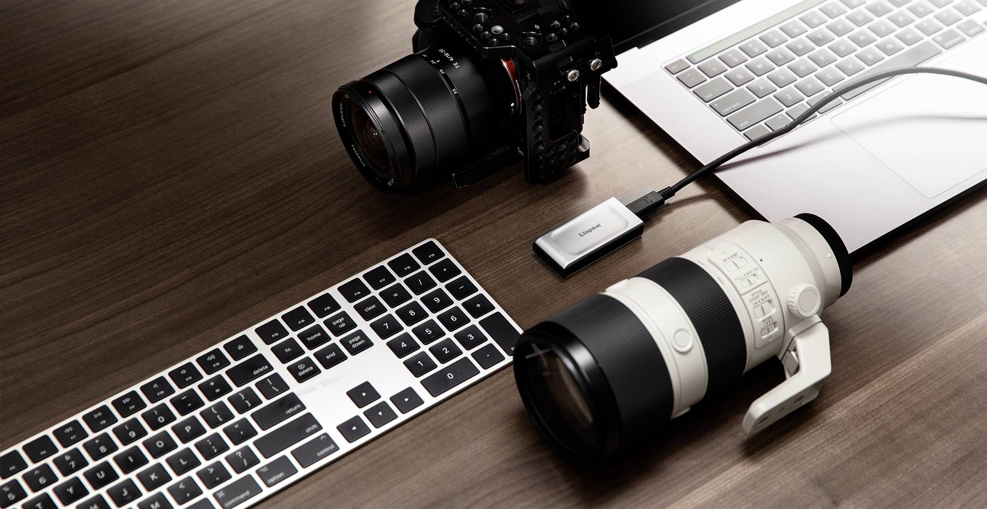 Zwei Kameras mit großen Objektiven liegen auf einem Holztisch neben einem Laptop und einer Tastatur. Eine Kamera ist per Kabel verbunden.