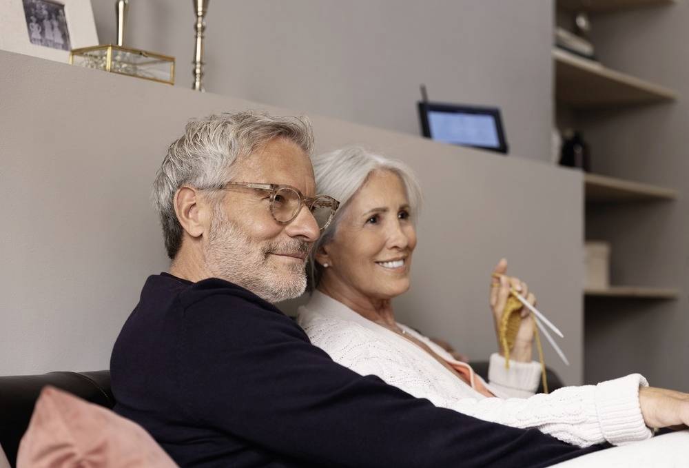 Ein älteres Paar sitzt lächelnd auf einem Sofa. Die Frau hält Stricknadeln und Wolle in der Hand. Der Raum ist gemütlich eingerichtet.