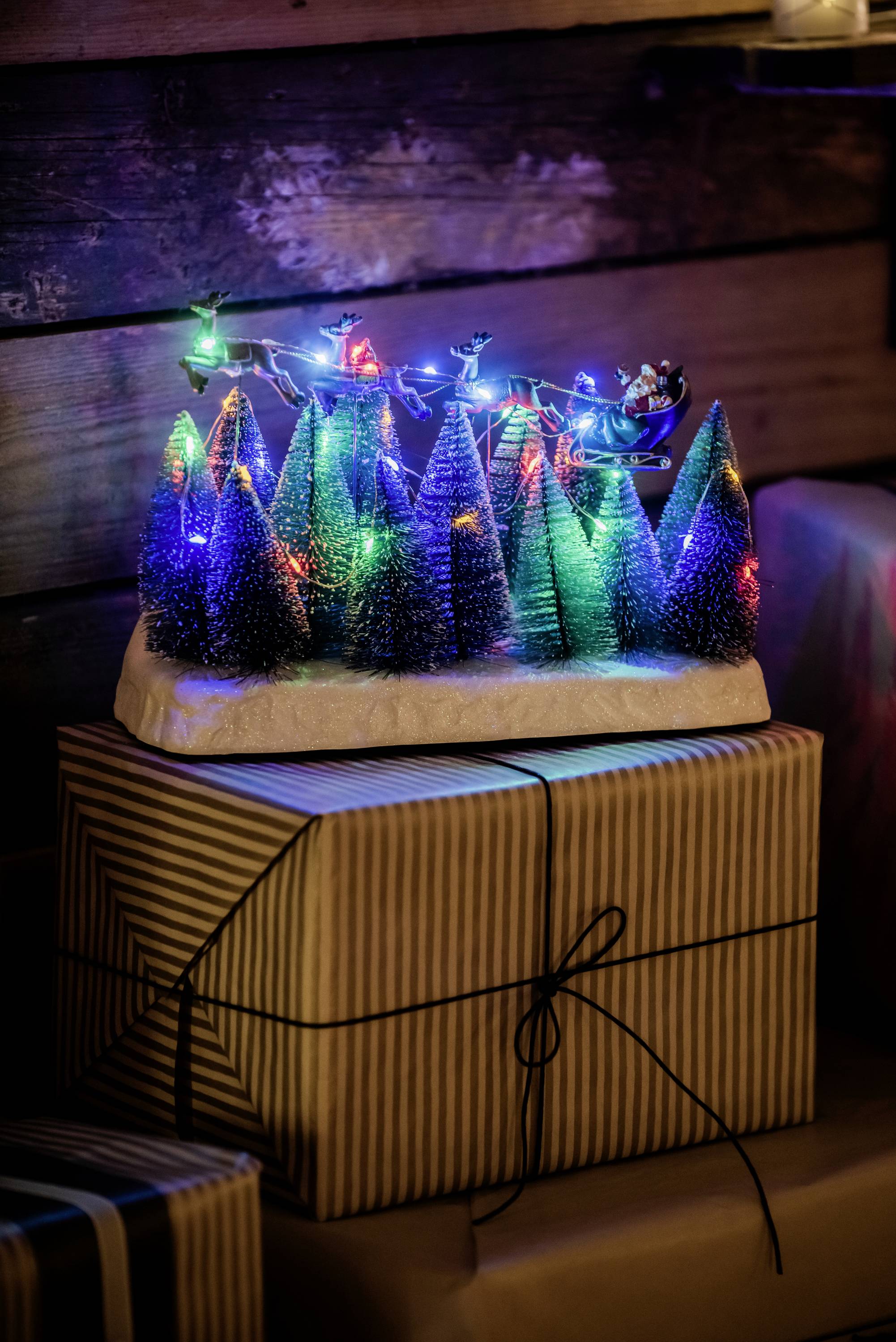 'Bunte LED-Lichterketten schmücken kleine Tannenbäume auf einem Geschenk, umgeben von weiteren Paketen. Festliche Weihnachtsdekoration.'
