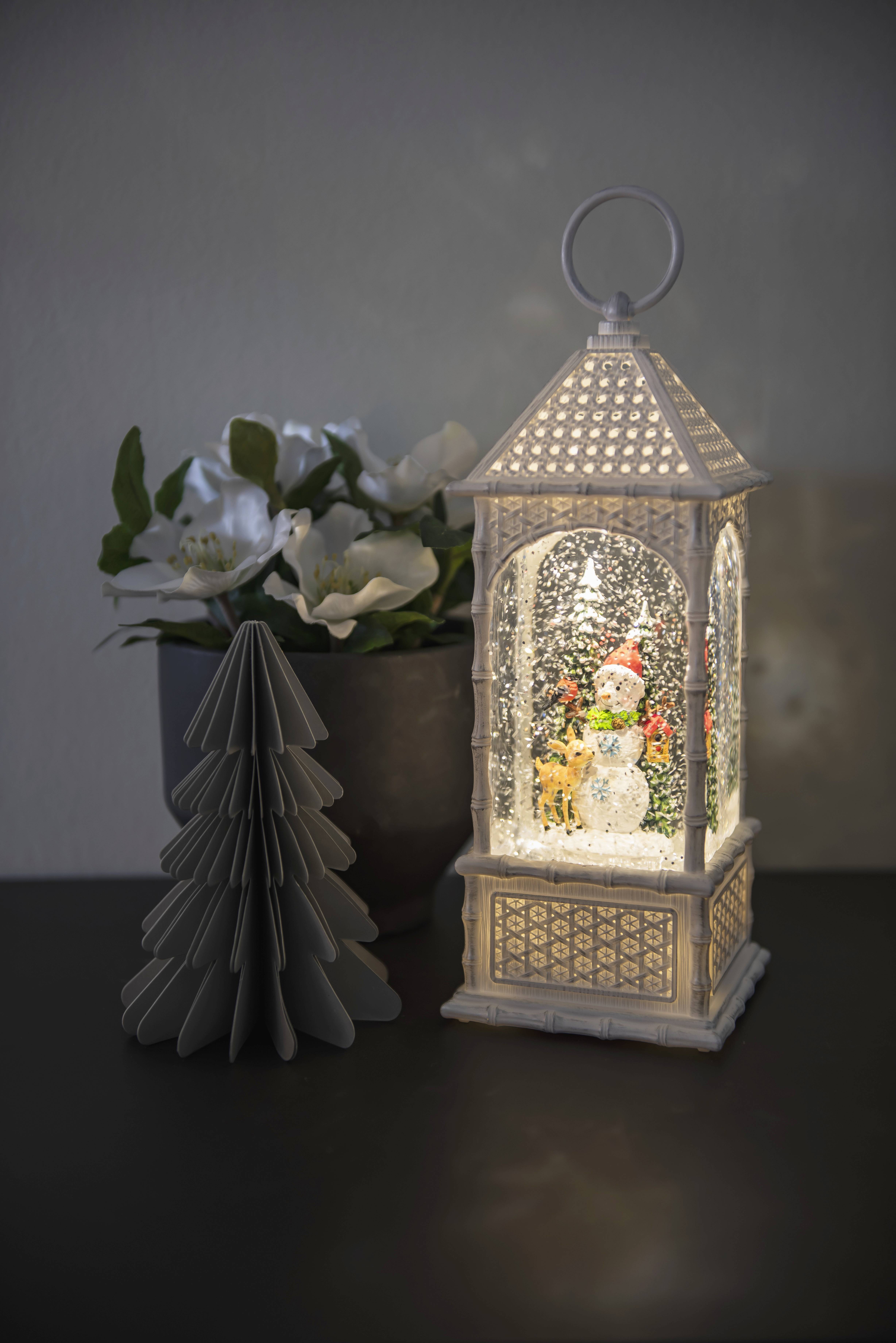 Eine beleuchtete Laterne mit Schneemann-Motiv, daneben weiße Blumen in einem Topf und ein kleiner, dekorativer Papierbaum.