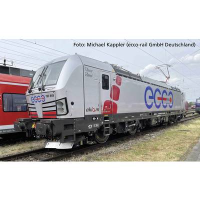 Graue Lokomotive mit rotem und blauem 'ecco' Logo. Seitliche Aufschrift 'Unser Hans' und 'akiem'. Befährt Bahngleise unter Stromleitungen.