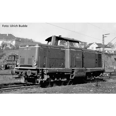 Eine alte schwarz-weiße Lokomotive auf Bahngleisen in einer städtischen Umgebung. Mehrere Gebäude sind im Hintergrund sichtbar.