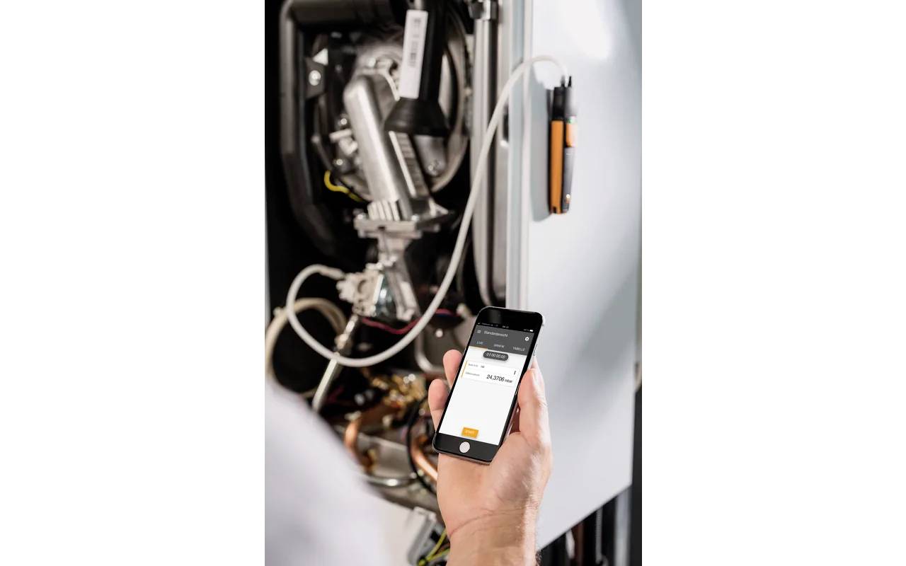 Eine Person hält ein Smartphone mit einer App, die eine Verbindung zu einem Heizgerät anzeigt. Im Hintergrund sind Teile des Heizgeräts sichtbar.
