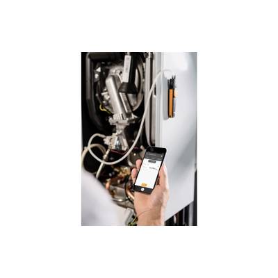 Eine Person hält ein Smartphone mit einer App, die eine Verbindung zu einem Heizgerät anzeigt. Im Hintergrund sind Teile des Heizgeräts sichtbar.