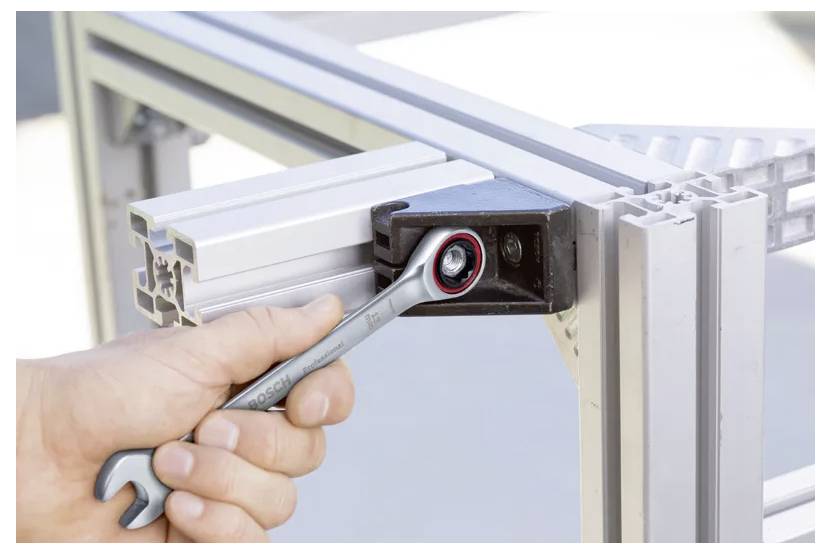 Eine Hand benutzt einen Schraubenschlüssel, um eine Schraube an einem Metallrahmen-Eckverbinder zu befestigen und zeigt dabei den Bau oder die Montage eines Strukturrahmens.