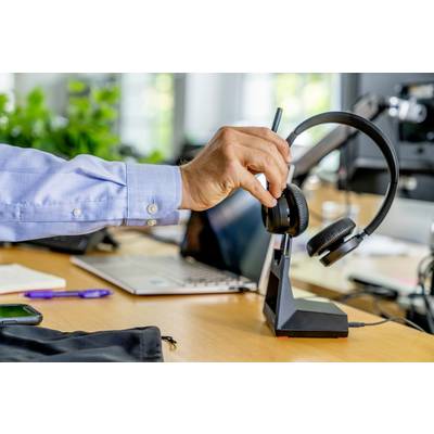 Eine Hand stellt ein Headset auf eine Dockingstation auf einem Schreibtisch ab, neben einem Laptop und einem Smartphone.