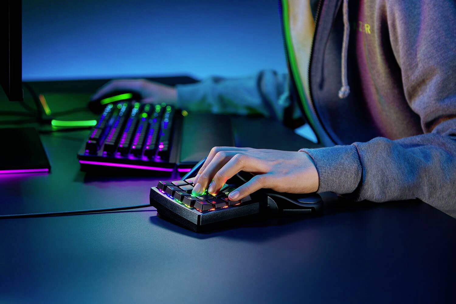 Person spielt an einem Computer mit einer beleuchteten Tastatur und einem Gamepad mit bunten Lichtern, in dunkler Umgebung.