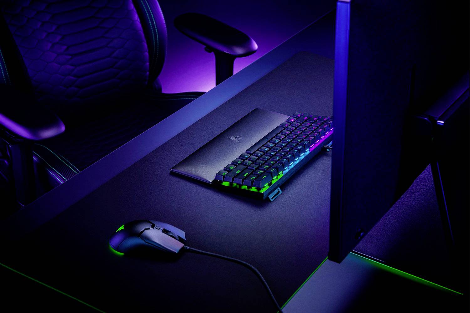 Schreibtisch mit Gaming-Tastatur, -Maus und -Monitor in dunkler Umgebung, beleuchtet von bunten LED-Lichtern. Links steht ein Bürostuhl.