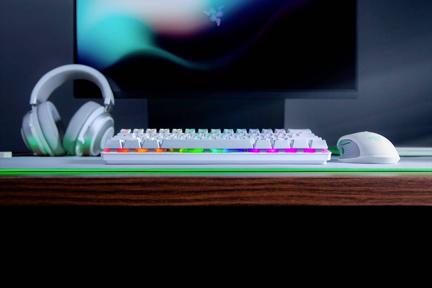 Eine beleuchtete Gaming-Tastatur, Maus und Kopfhörer auf einem Holzschreibtisch vor einem unscharfen Bildschirm.